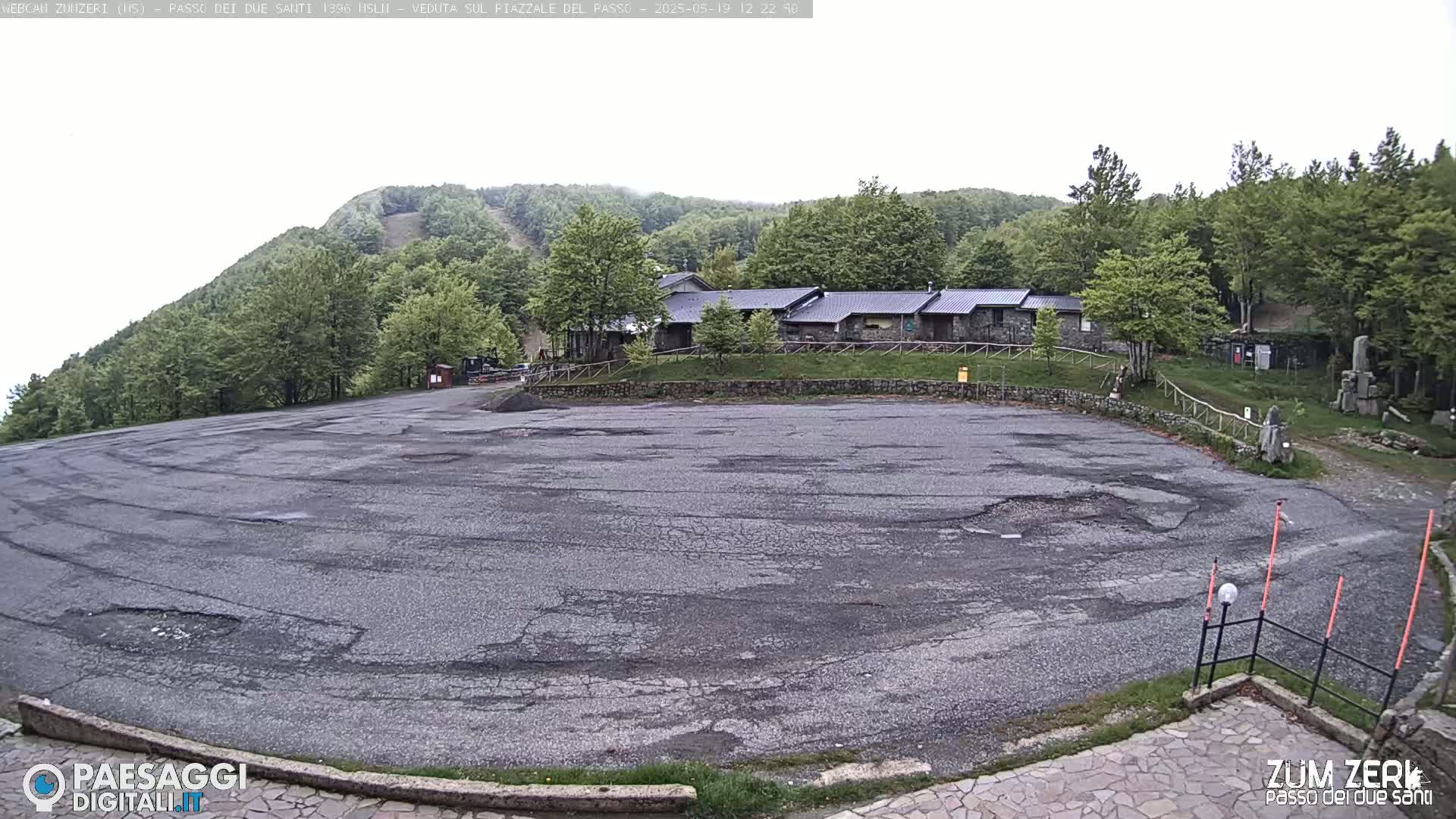 A large, cracked asphalt parking lot sits before a cluster of stone buildings nestled among green hills under an overcast sky.