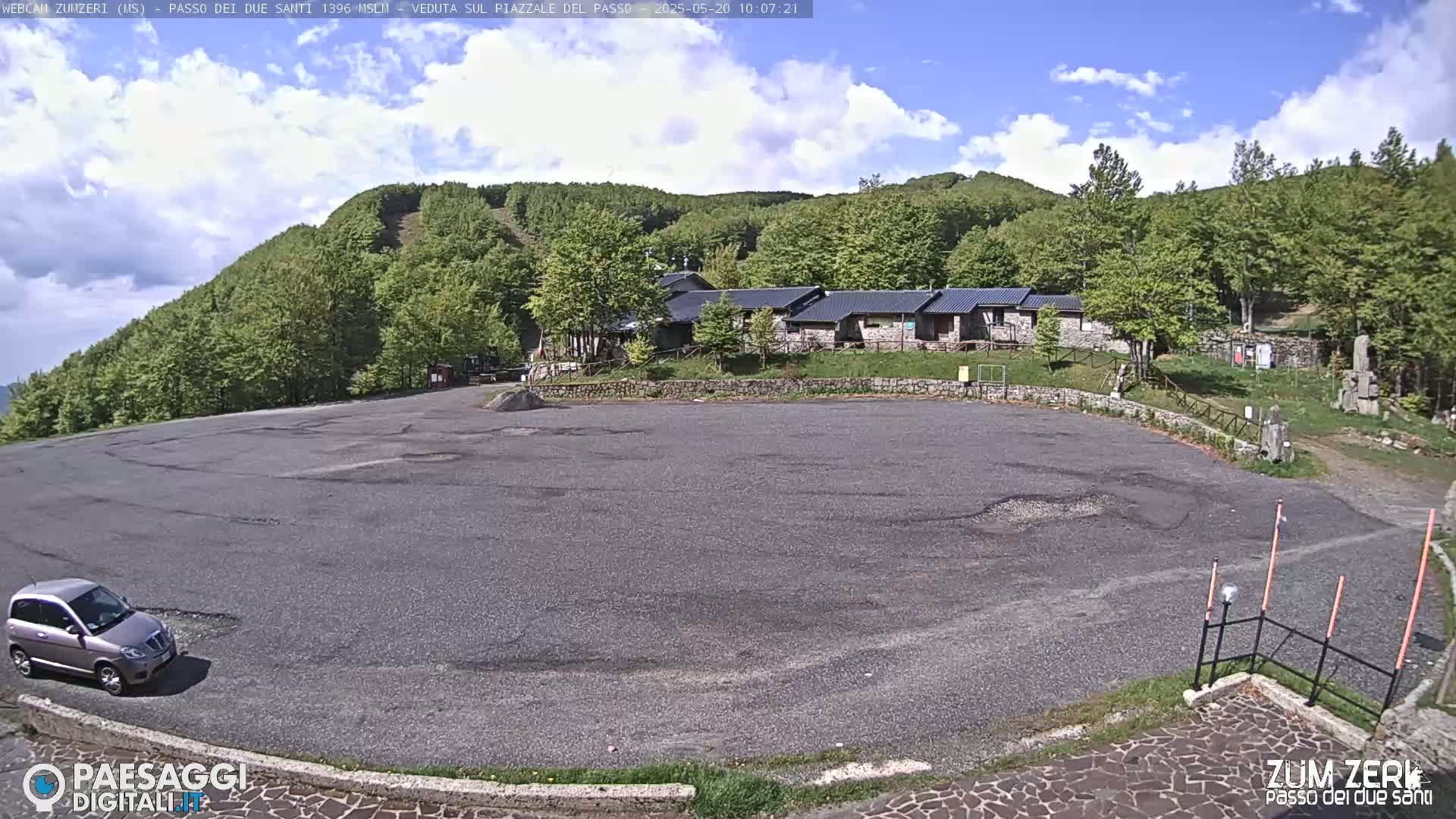 A mostly empty asphalt parking lot sits below a cluster of buildings nestled in a hillside of lush green trees under a partly cloudy sky.