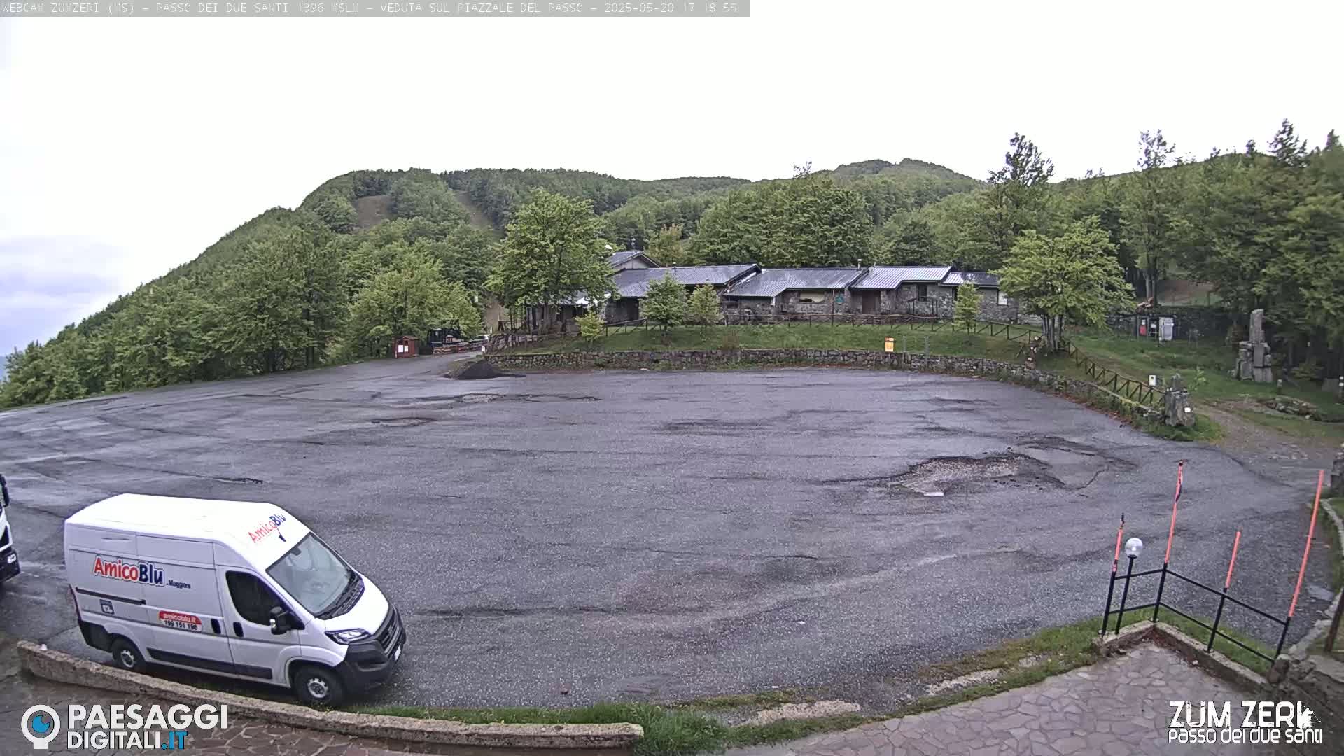A mostly empty asphalt parking lot sits in front of a cluster of buildings nestled among green hills under an overcast sky.