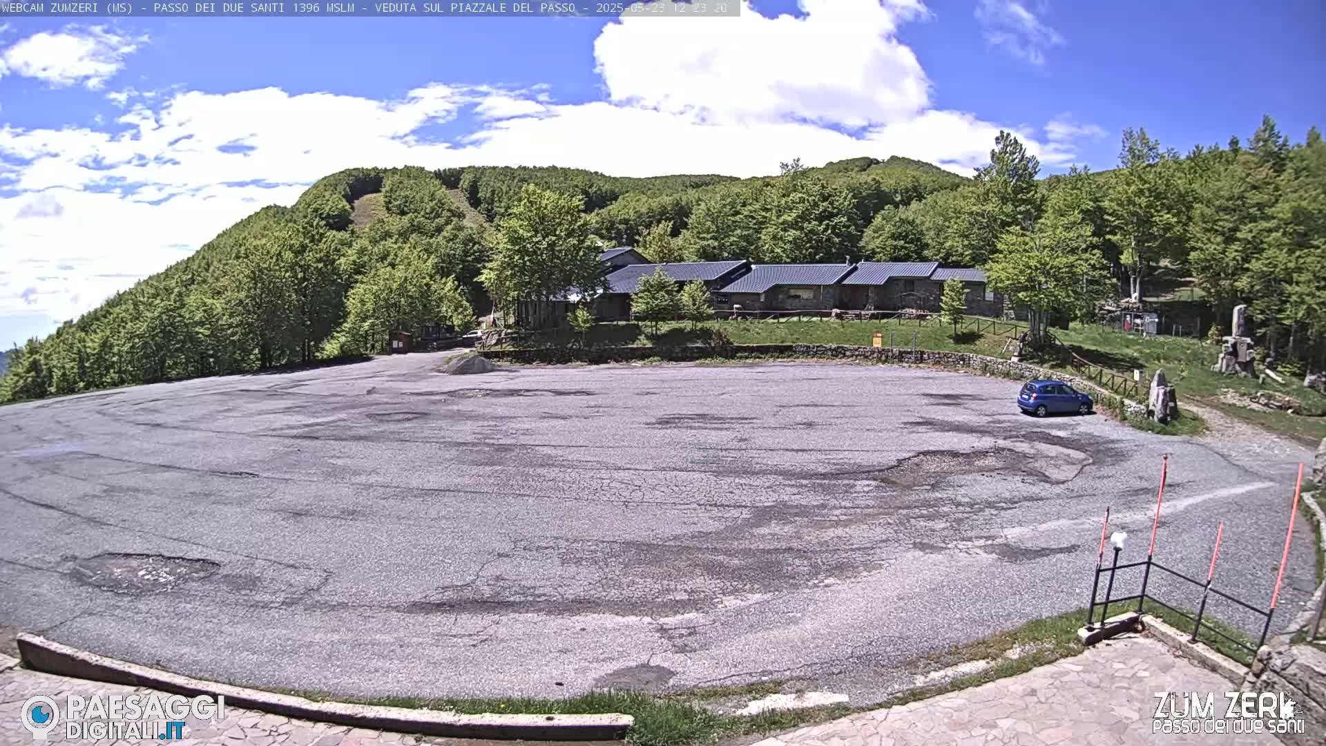 A mostly empty asphalt parking lot sits below a row of buildings nestled among a verdant hillside under a partly cloudy sky.