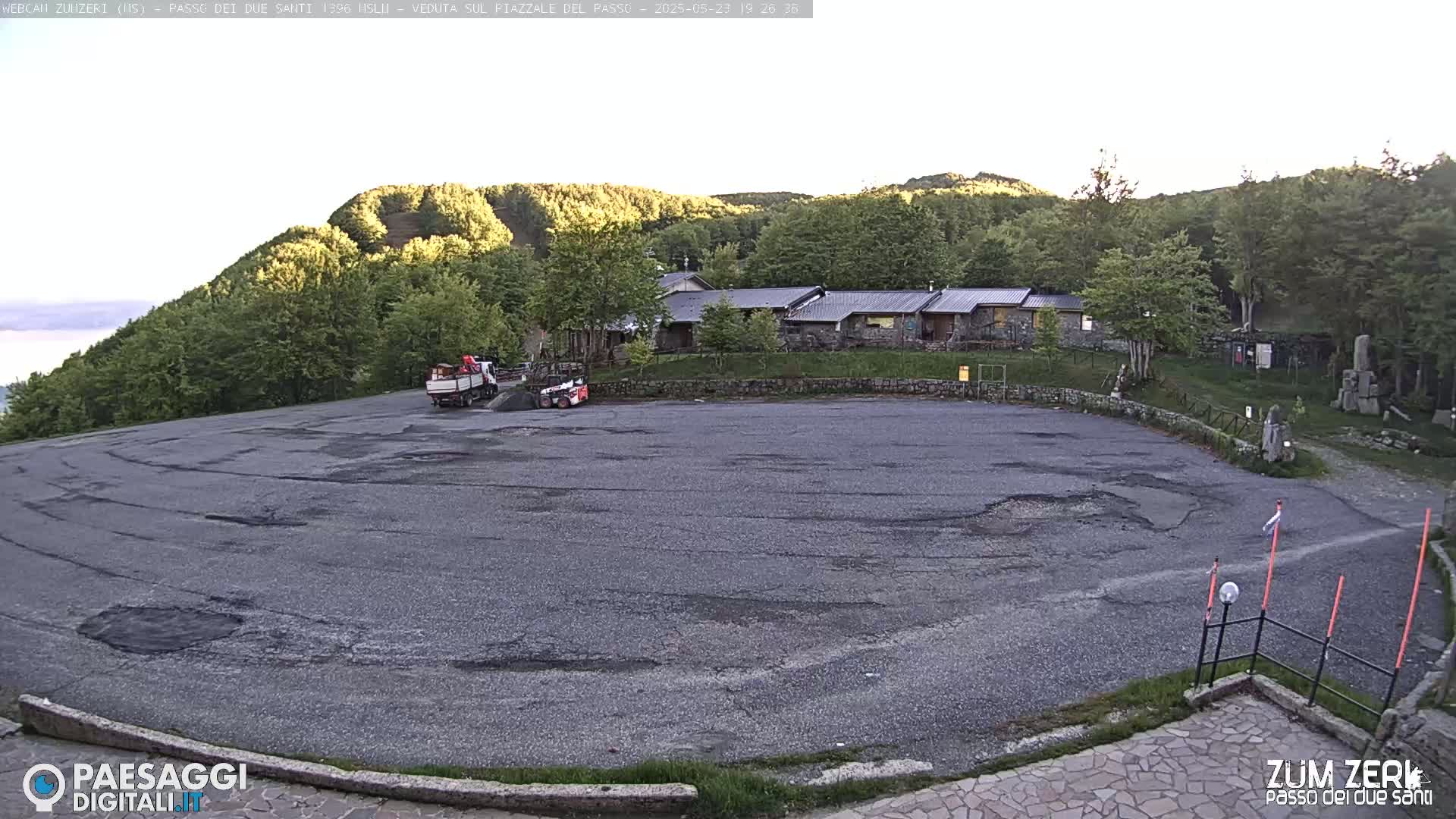 A large, empty asphalt parking lot sits below a cluster of stone buildings nestled amongst green hills under a clear, light sky.