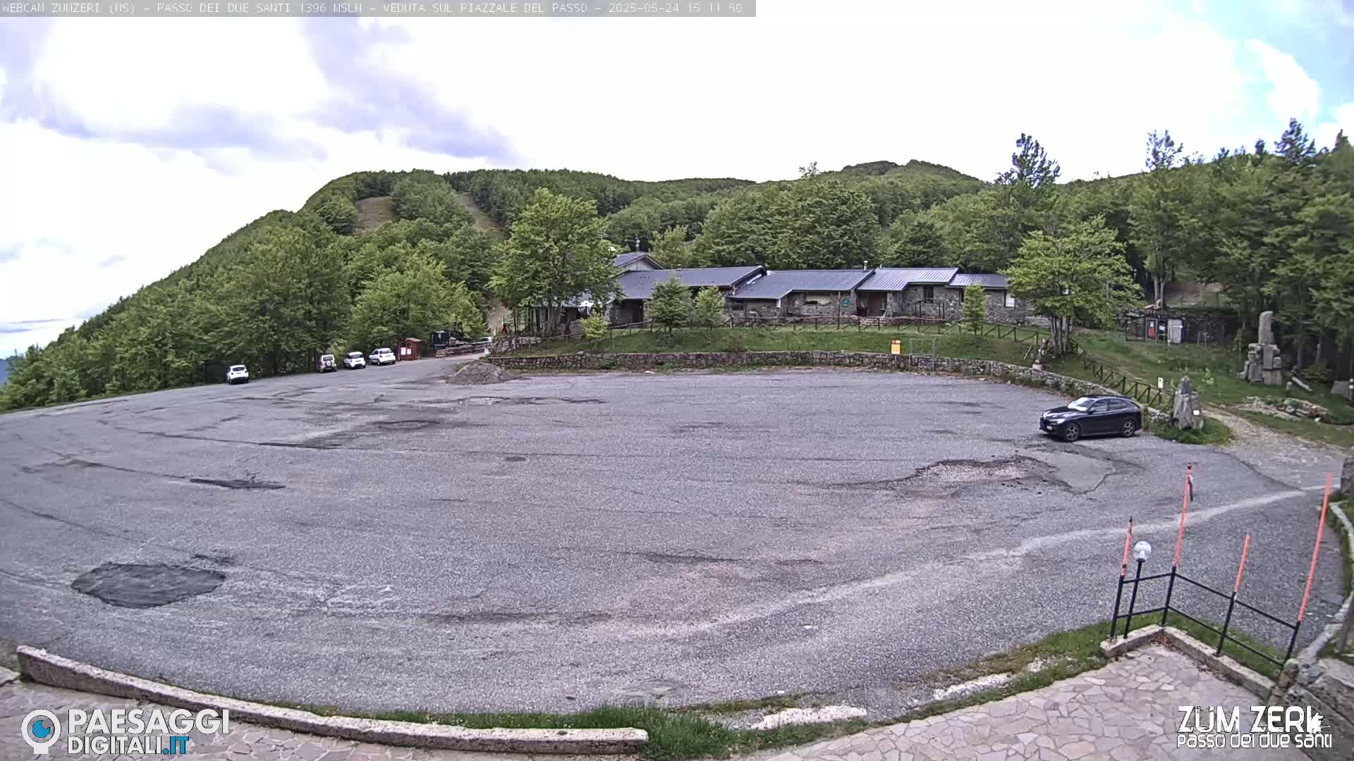 A mostly empty asphalt parking lot sits in front of a stone building nestled among a lush green hillside under a partly cloudy sky.