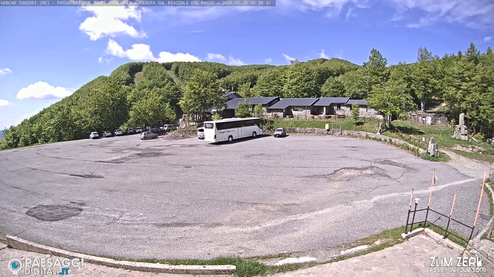 A mostly empty parking lot with a large bus and several cars is situated in front of a building complex nestled amongst a lush, green, hilly landscape under a mostly sunny sky.