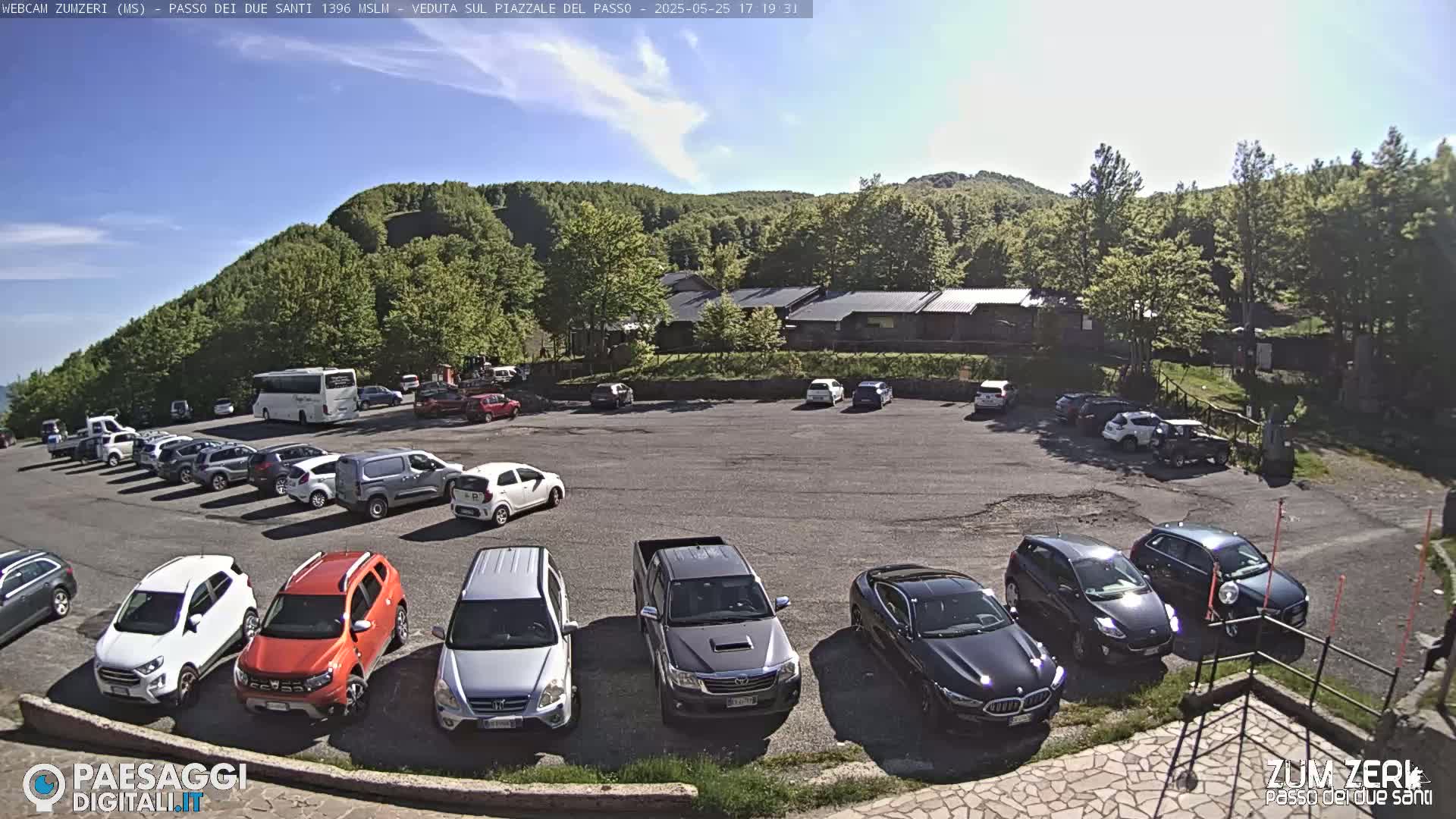 A parking lot filled with cars and a bus is situated below a building, against a backdrop of a green, tree-covered hill under a mostly clear sky.