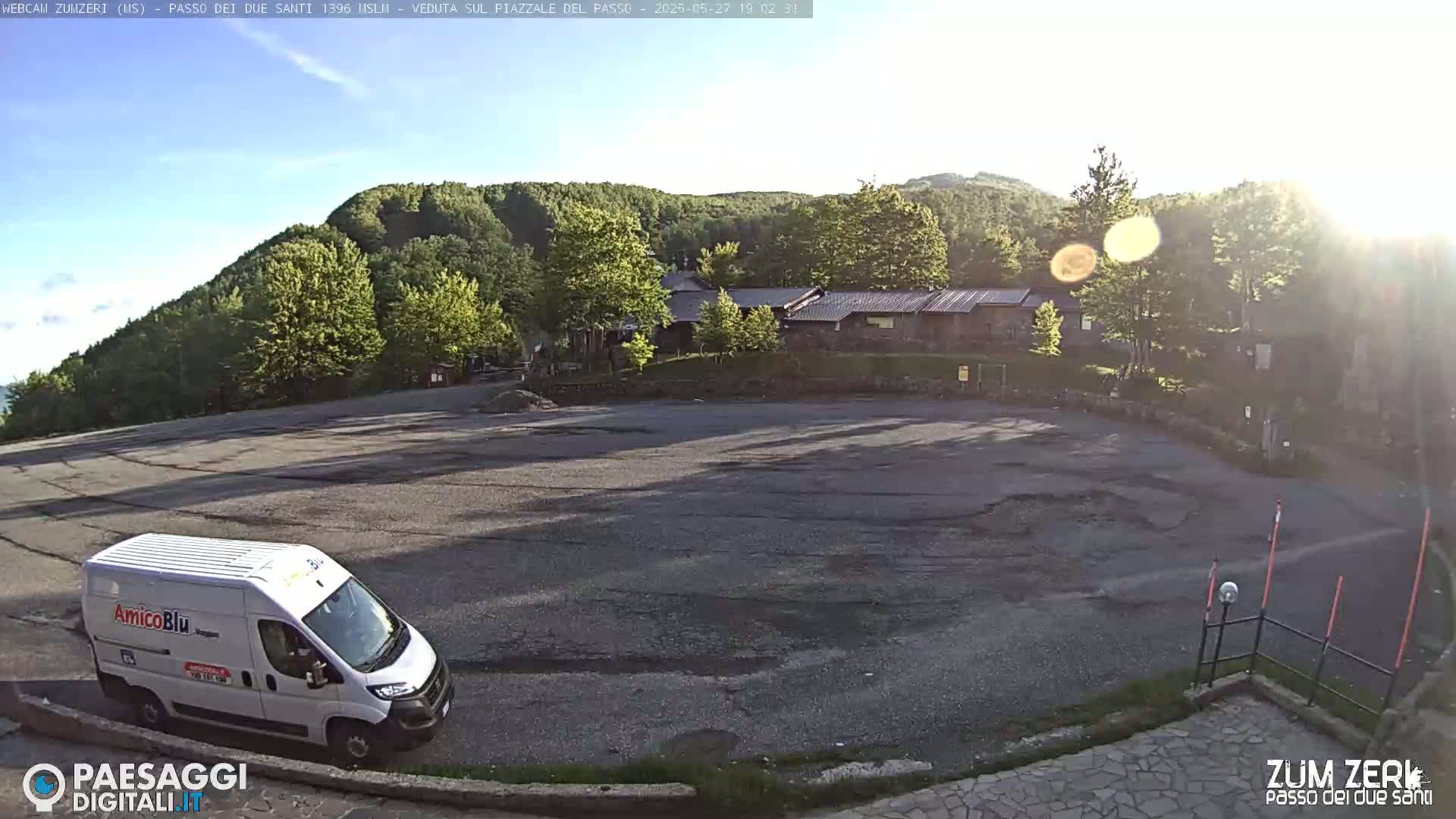 A white van is parked in a large, empty asphalt lot surrounded by trees and a building under a mostly sunny sky.