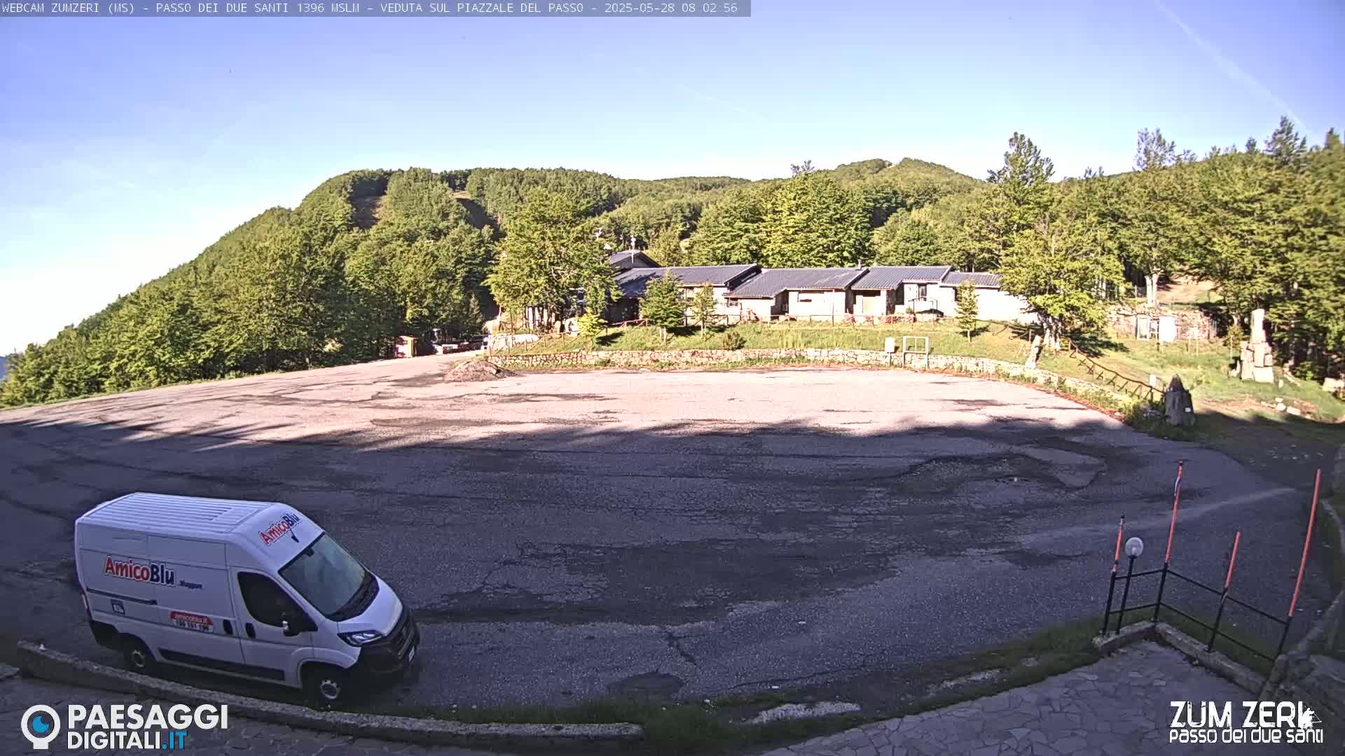 A white van is parked in a cracked asphalt parking lot, overlooking a cluster of buildings nestled in a lush green hillside under a clear blue sky.