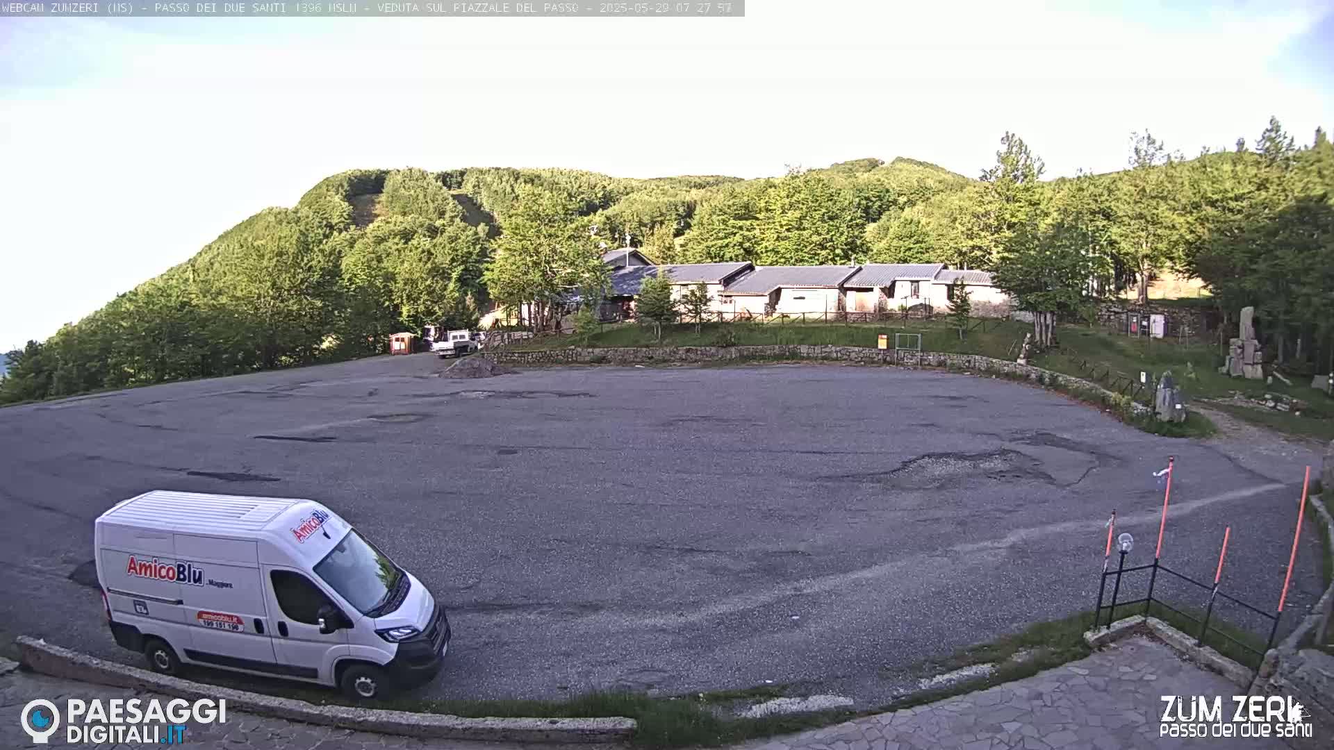 A mostly empty asphalt parking lot sits before a row of buildings nestled in a hillside of green trees under a partly cloudy sky.