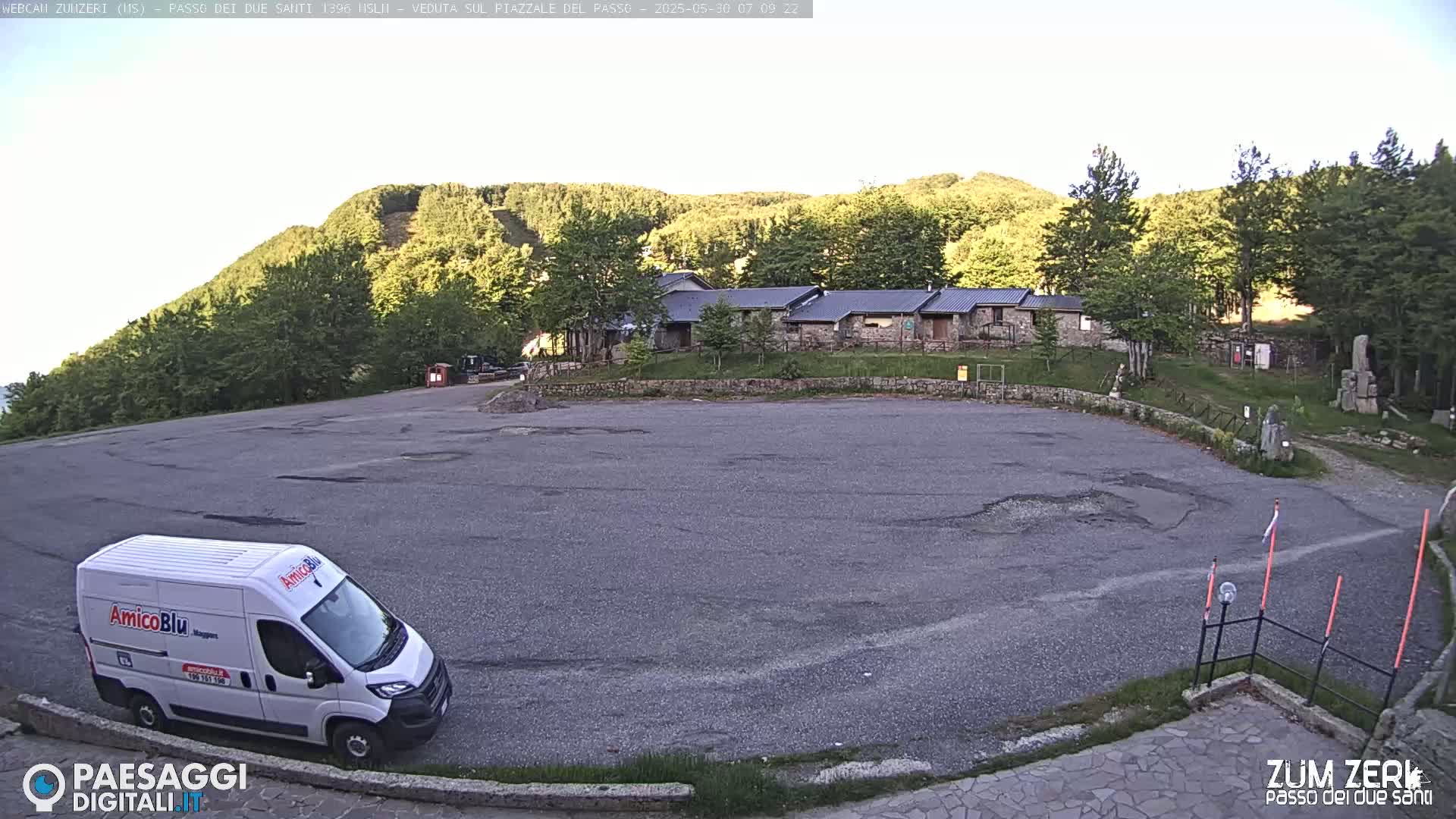 A mostly empty asphalt parking lot sits in front of a low stone building, with a white van parked to the left, all nestled among green hills under a clear, light-blue sky.