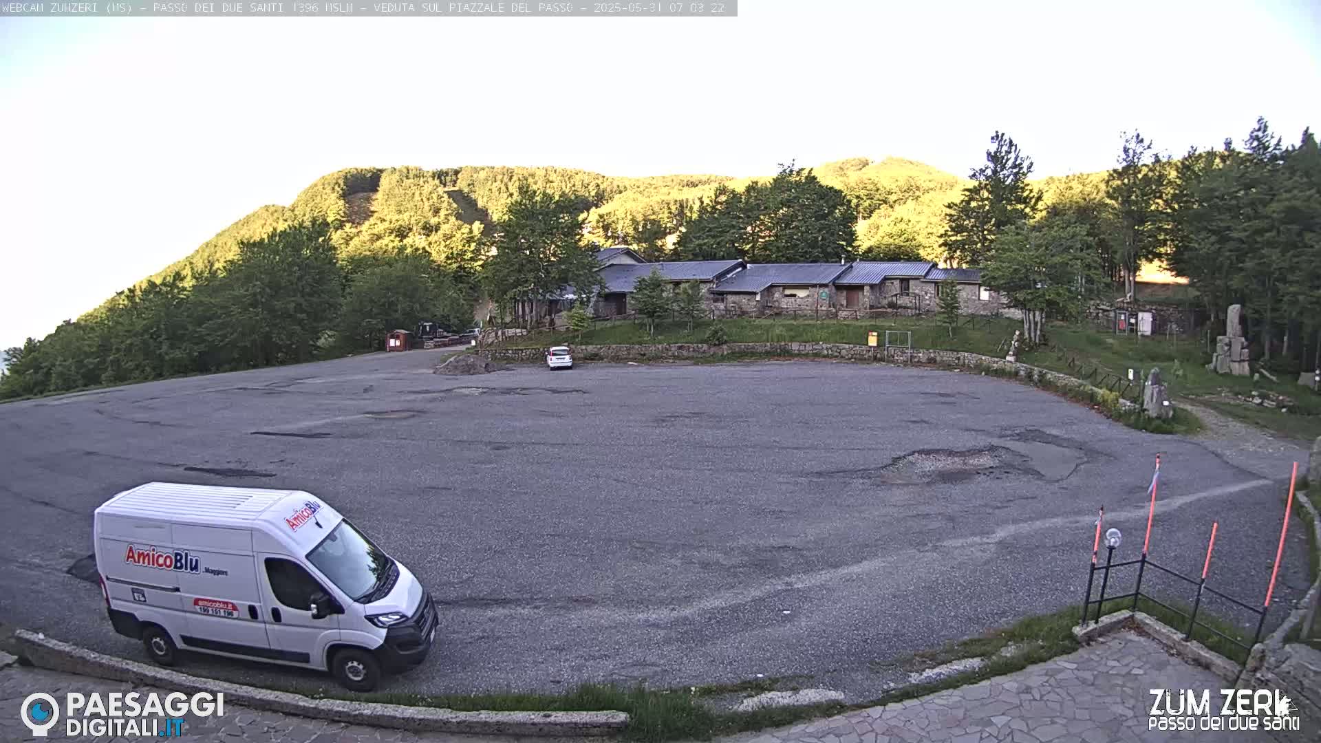 A mostly empty asphalt parking lot sits before a stone building complex nestled in a mountainside forest under a clear, bright sky.