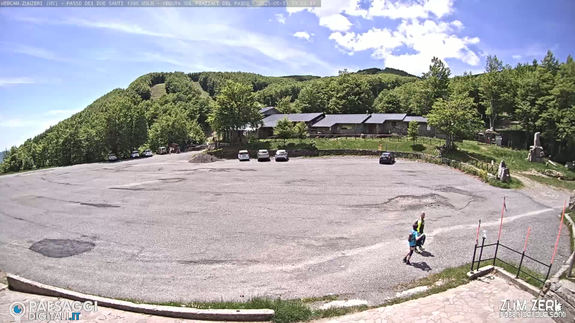 A mostly empty asphalt parking lot sits below a building complex nestled in a green, forested hillside under a partly cloudy sky.