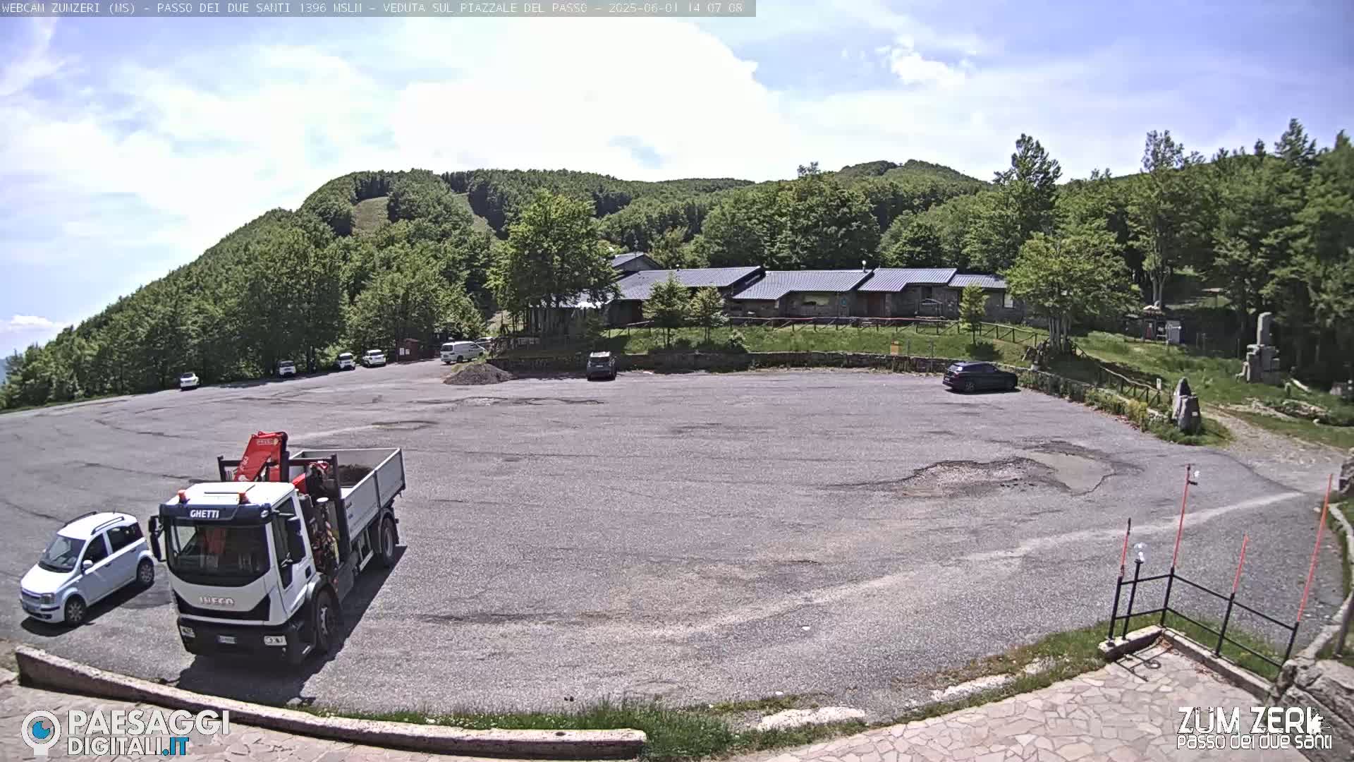 A mostly empty asphalt parking lot sits in front of a building complex nestled within a green, wooded hillside under a partly cloudy sky.