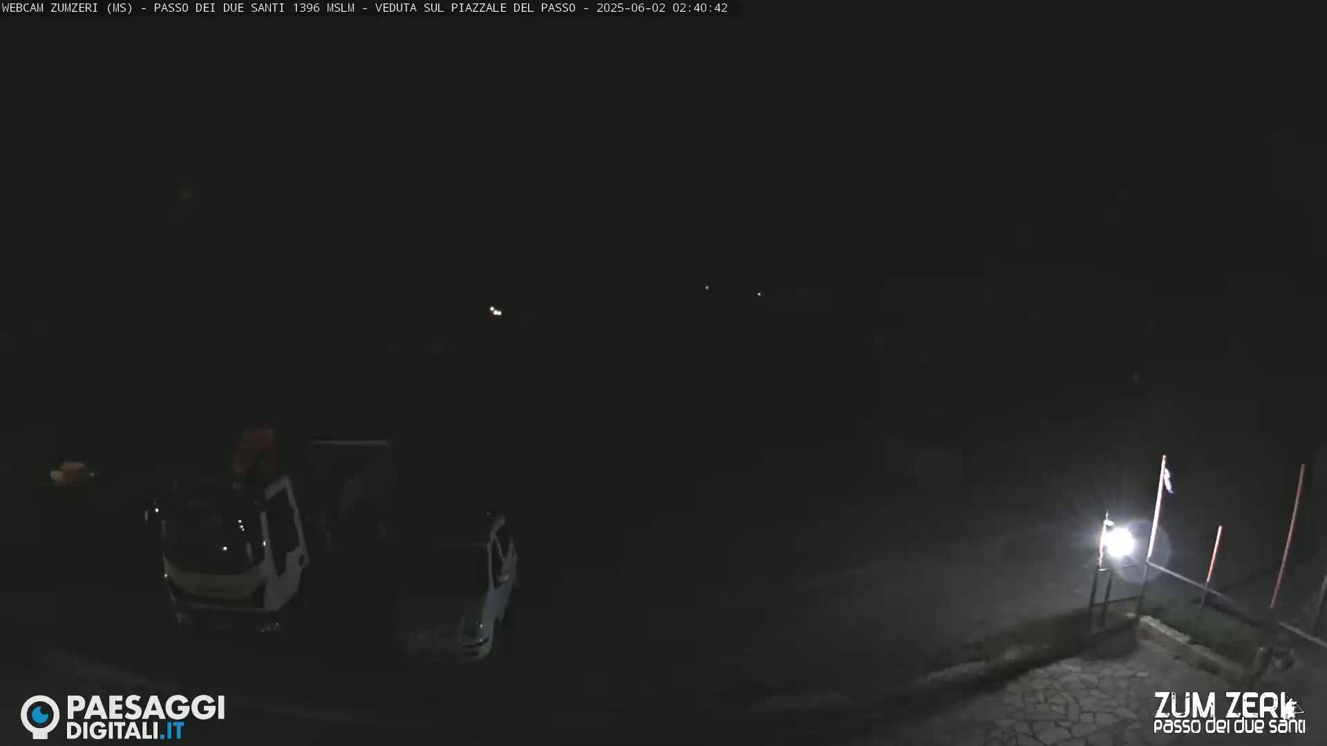 At night, two vehicles are parked in a dimly lit area, with a single light illuminating a nearby paved surface.