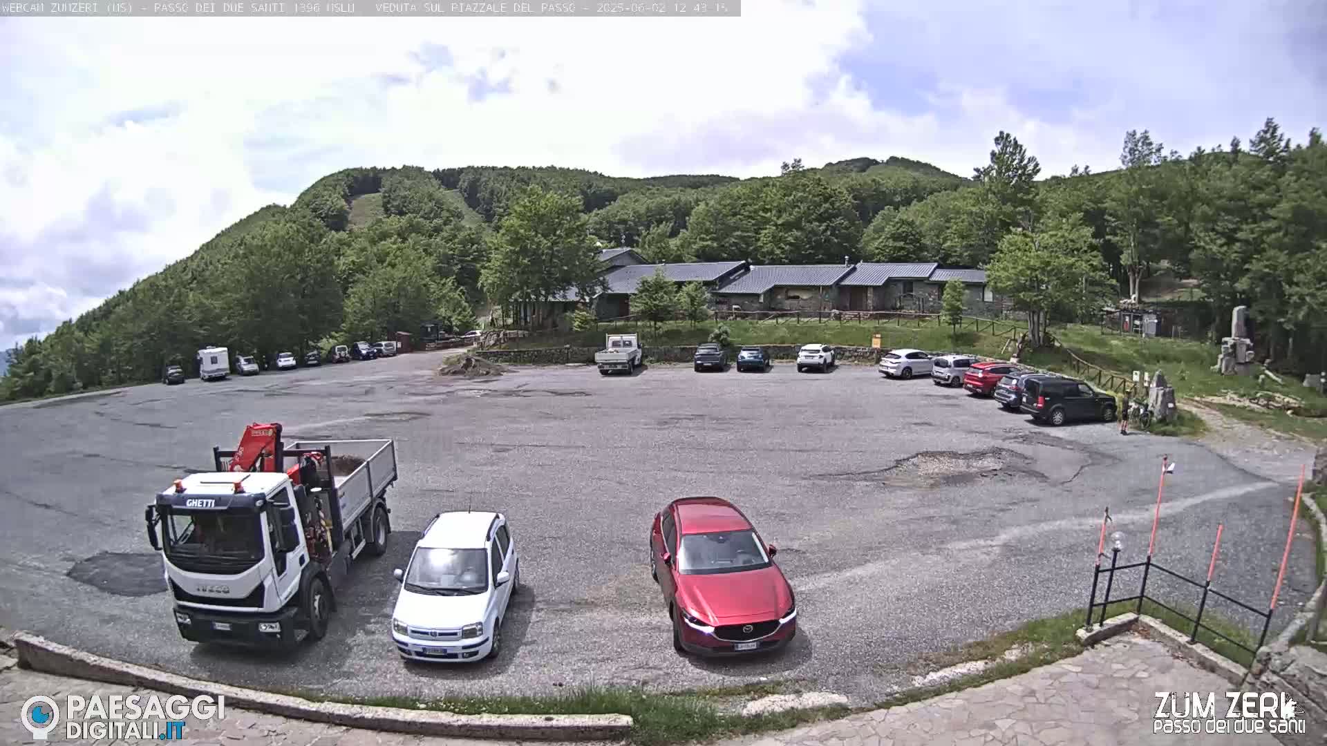 A parking lot with several cars and a truck is situated in front of a building,  surrounded by a lush green mountainside under a partly cloudy sky.
