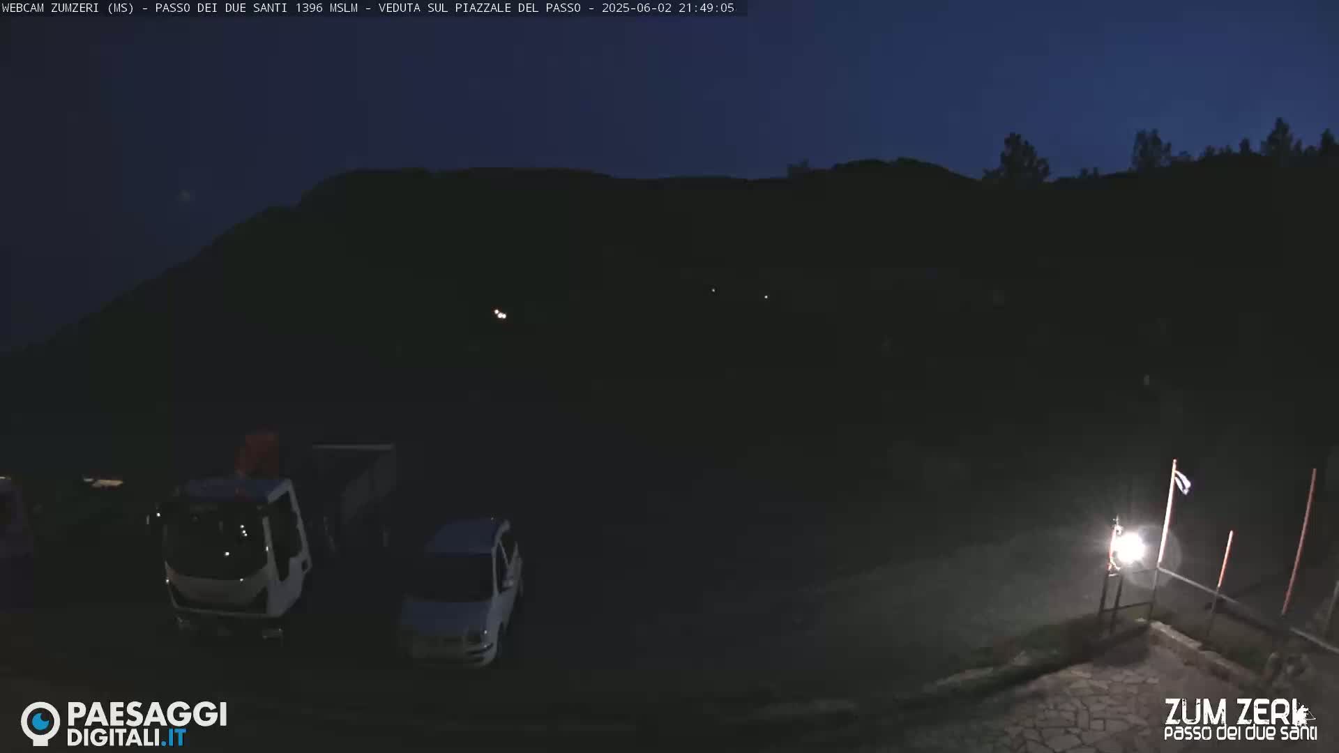 At night, under a dark sky, a few vehicles are parked in a dimly lit area near a hill.