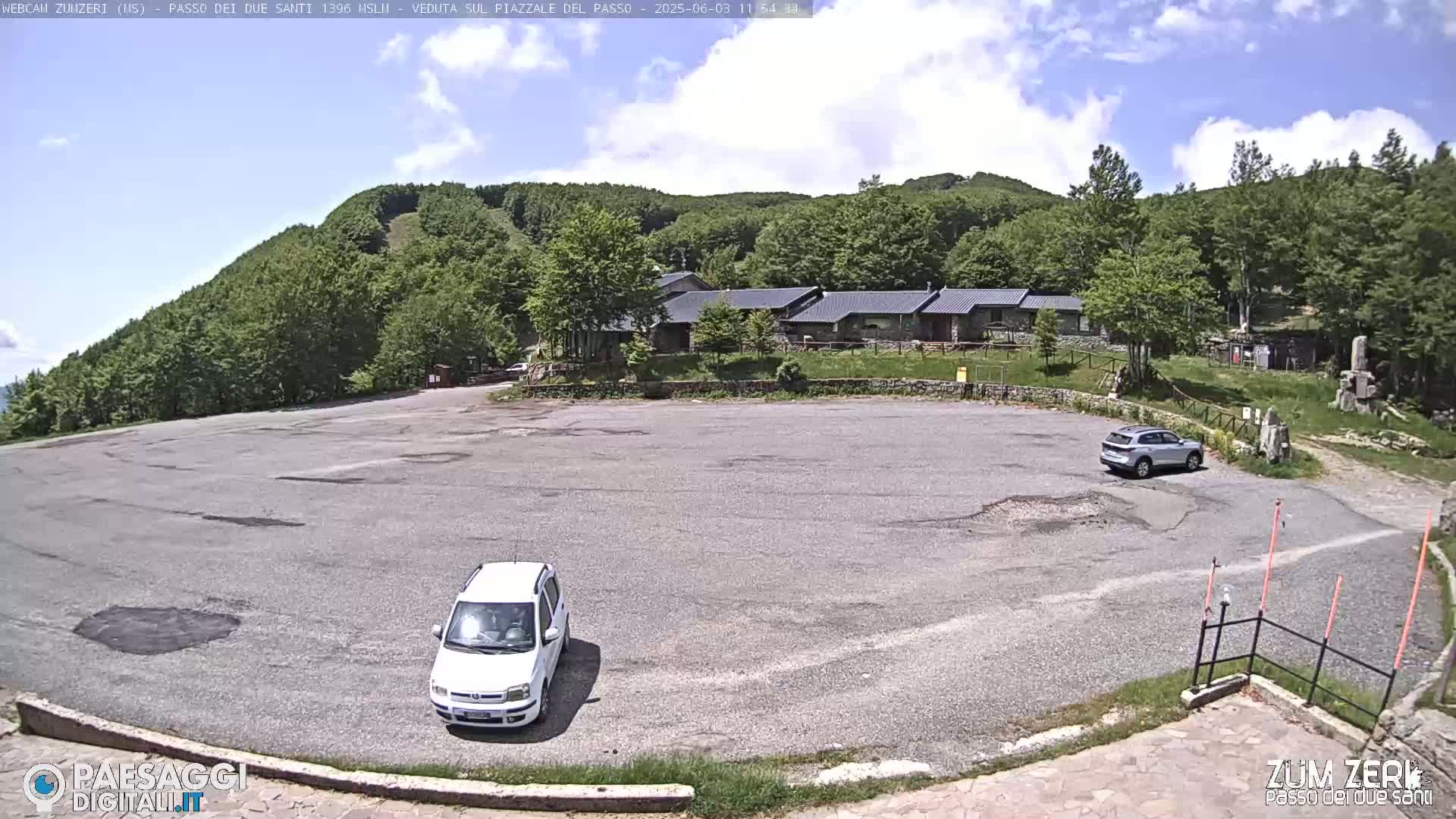 A mostly empty asphalt parking lot sits in front of a low building complex, surrounded by a lush green hill covered in trees under a partly cloudy sky.