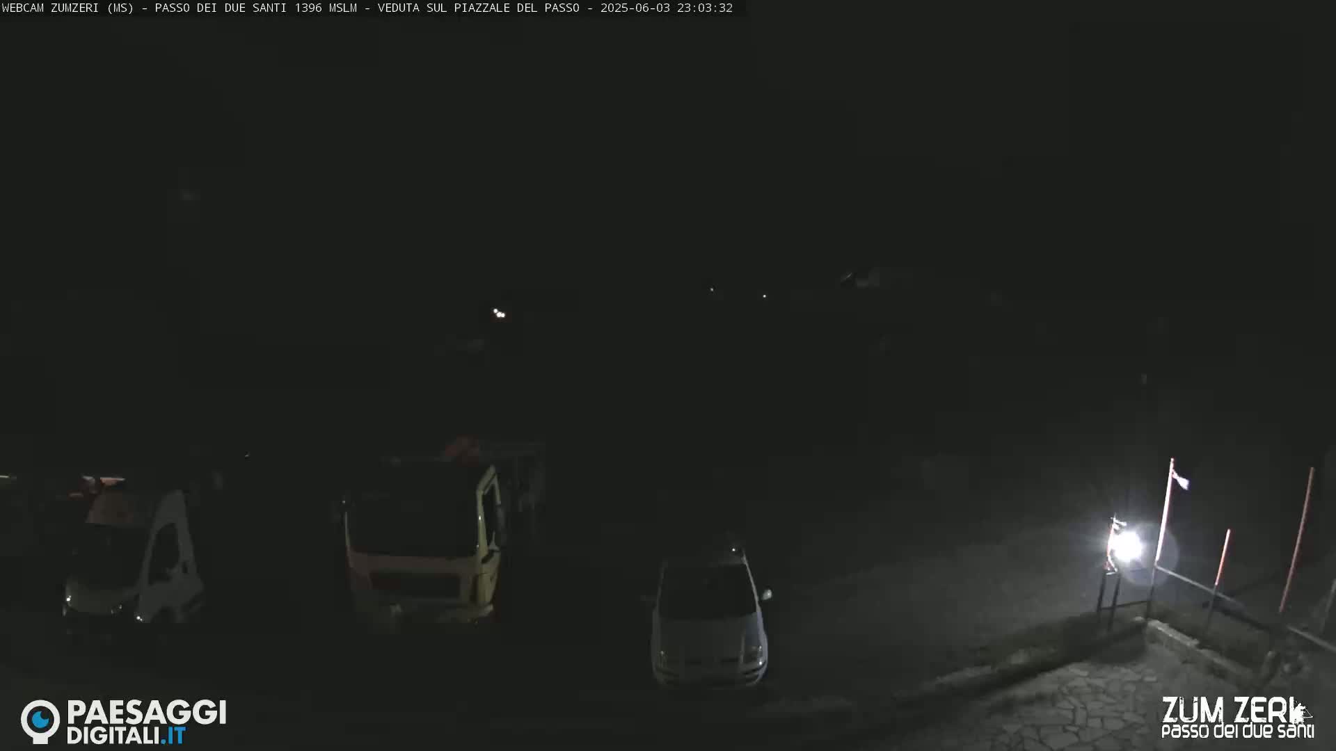 A dark, nighttime scene shows a parking lot with a few vehicles, illuminated by a single light.