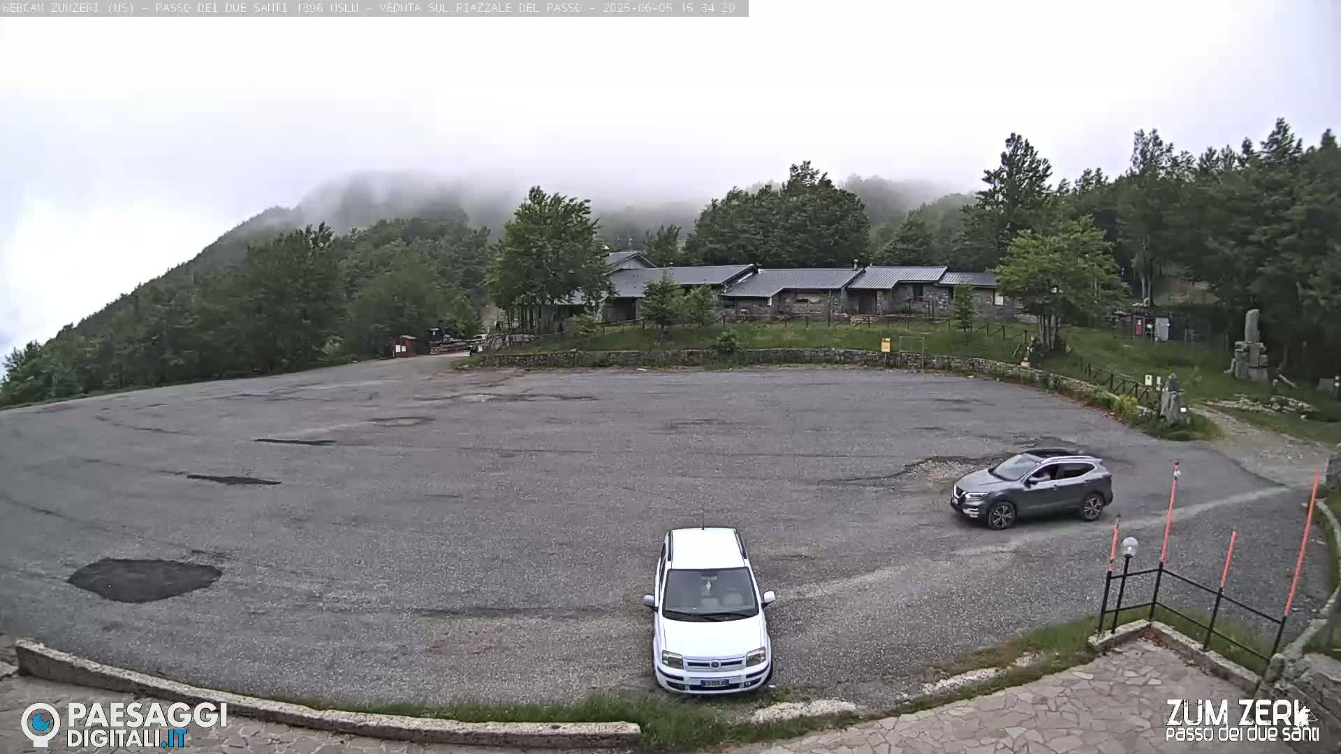 A mostly empty asphalt parking lot sits below a building complex nestled in a foggy, green mountainside.