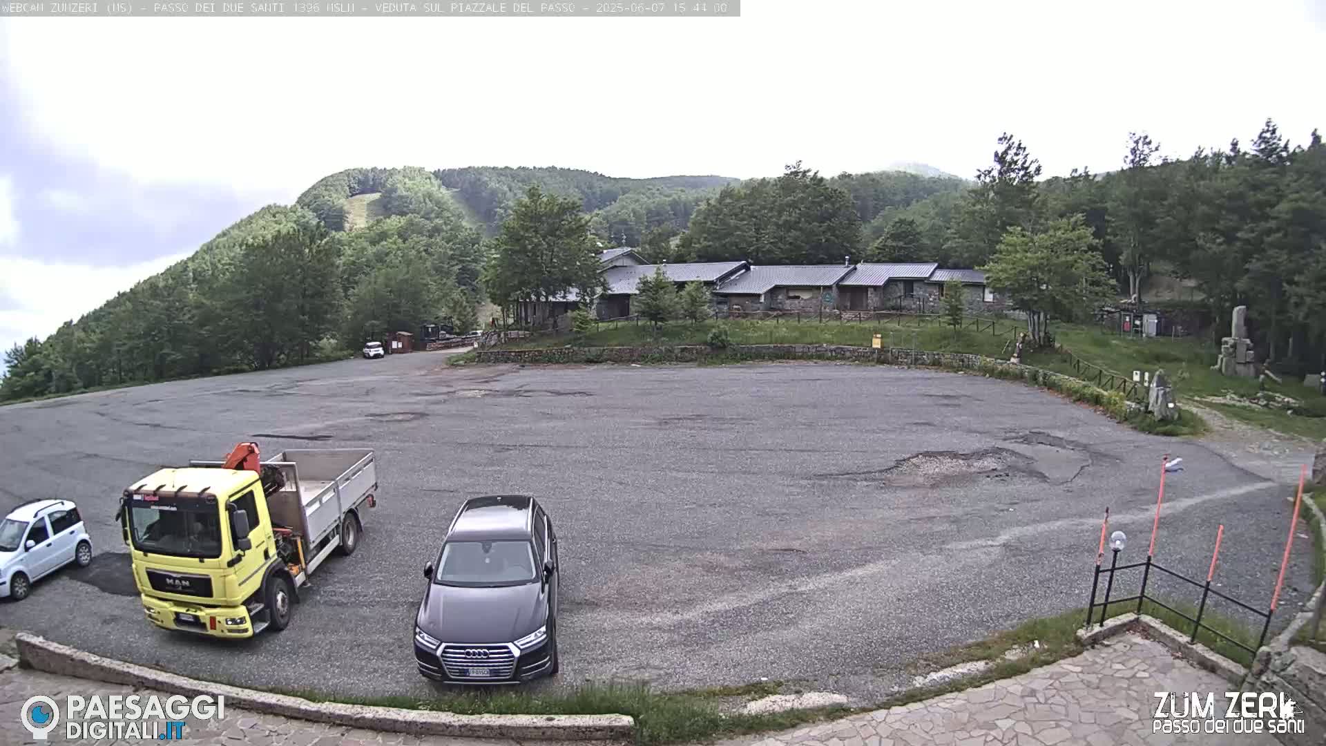 A large empty asphalt parking lot sits before a group of buildings nestled among a lush green hillside under an overcast sky.