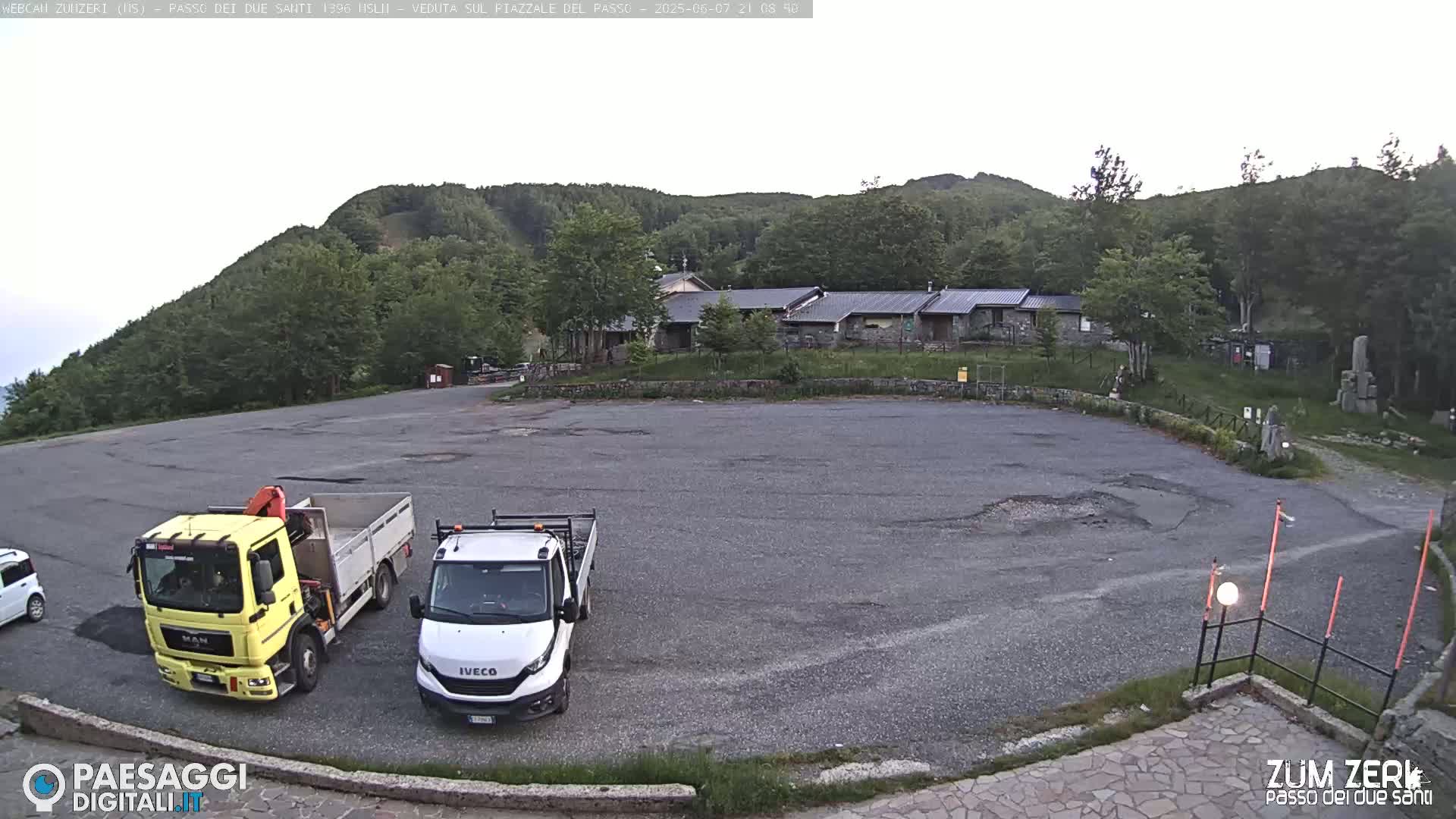 A mostly empty asphalt parking lot with two trucks and a car, situated in front of a cluster of buildings nestled within a green, hilly landscape under a mostly cloudy sky.