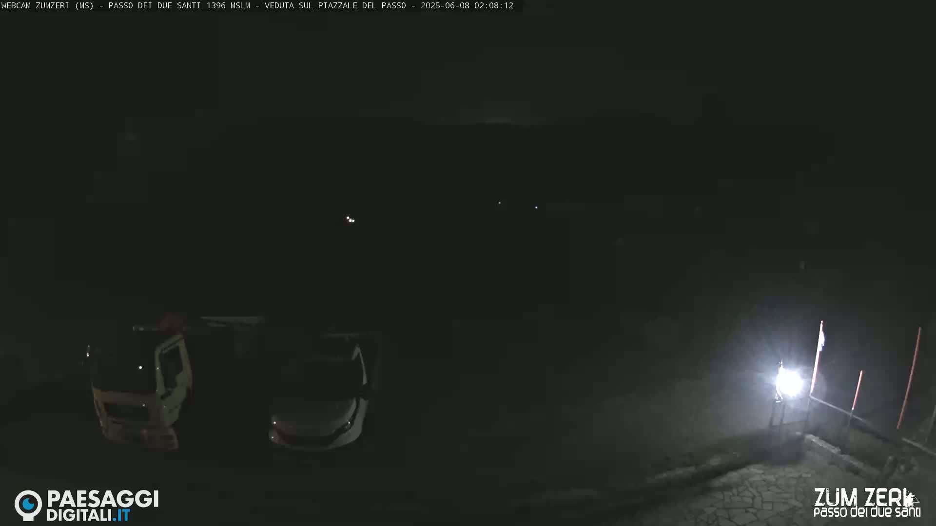 A dark, nighttime scene shows a truck and car parked in a mostly empty lot, illuminated by a bright light in the lower right corner.