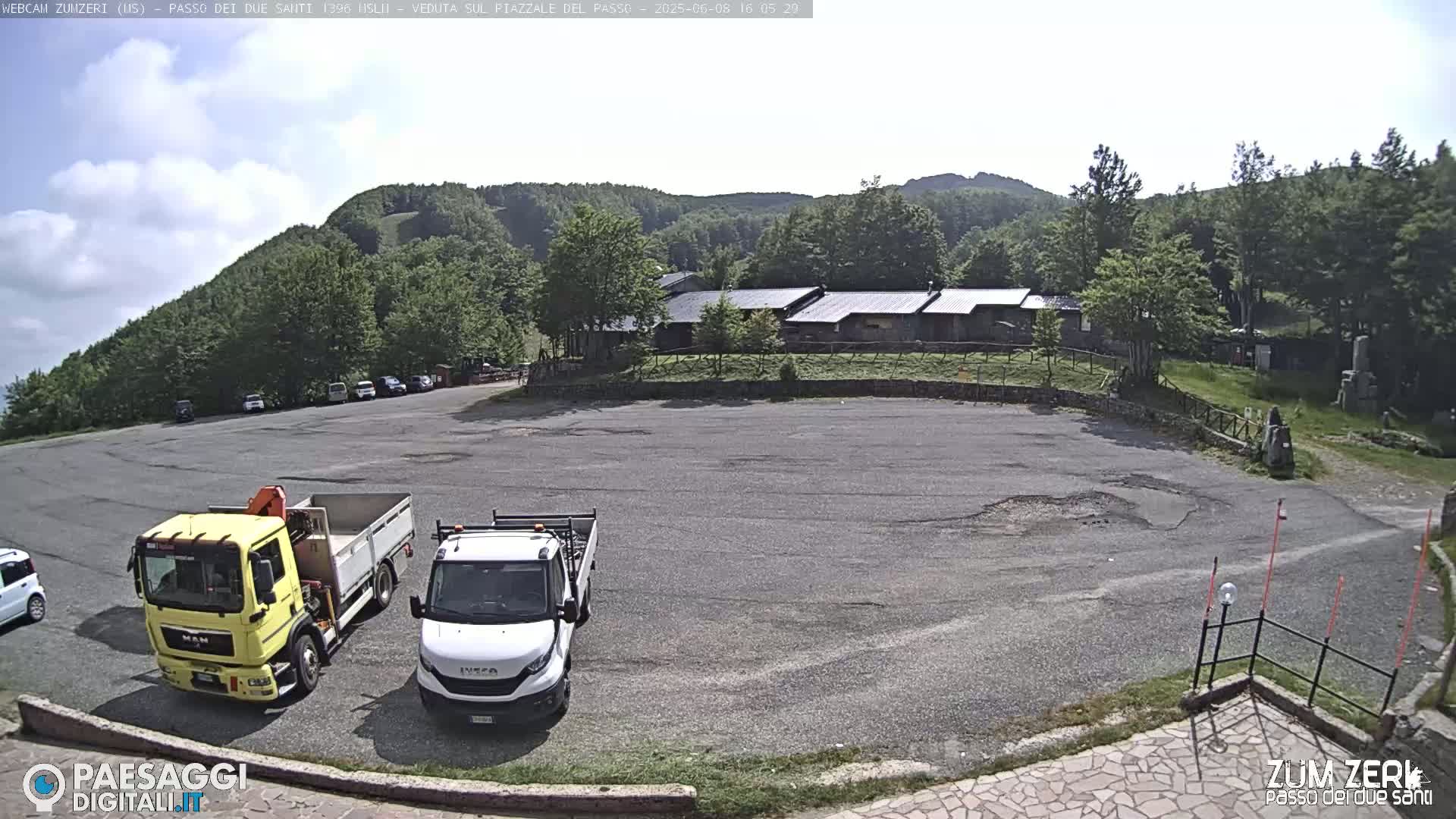 A mostly empty asphalt parking lot is situated below a group of low buildings near a mountainside under a partly cloudy sky.