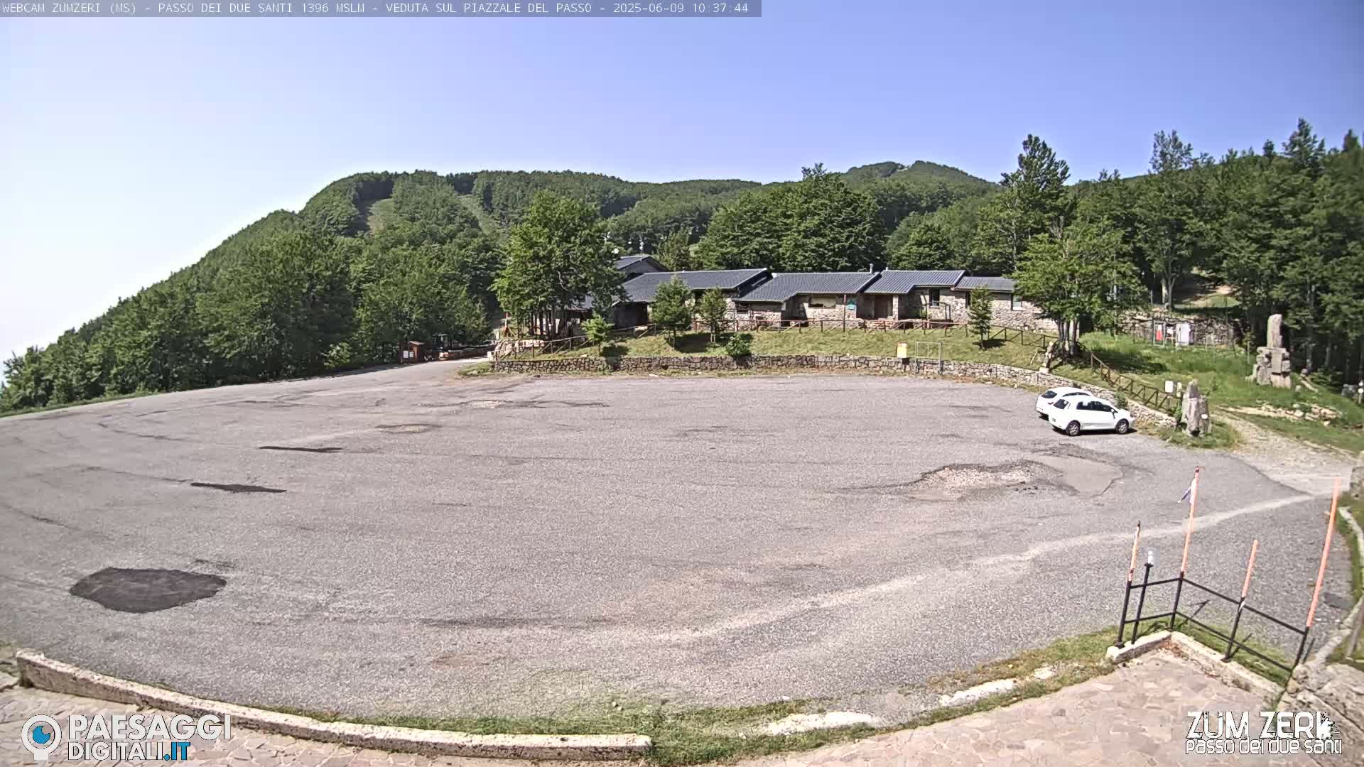 A mostly empty asphalt parking lot sits before a low stone building complex nestled against a hillside covered in lush green forest under a clear blue sky.