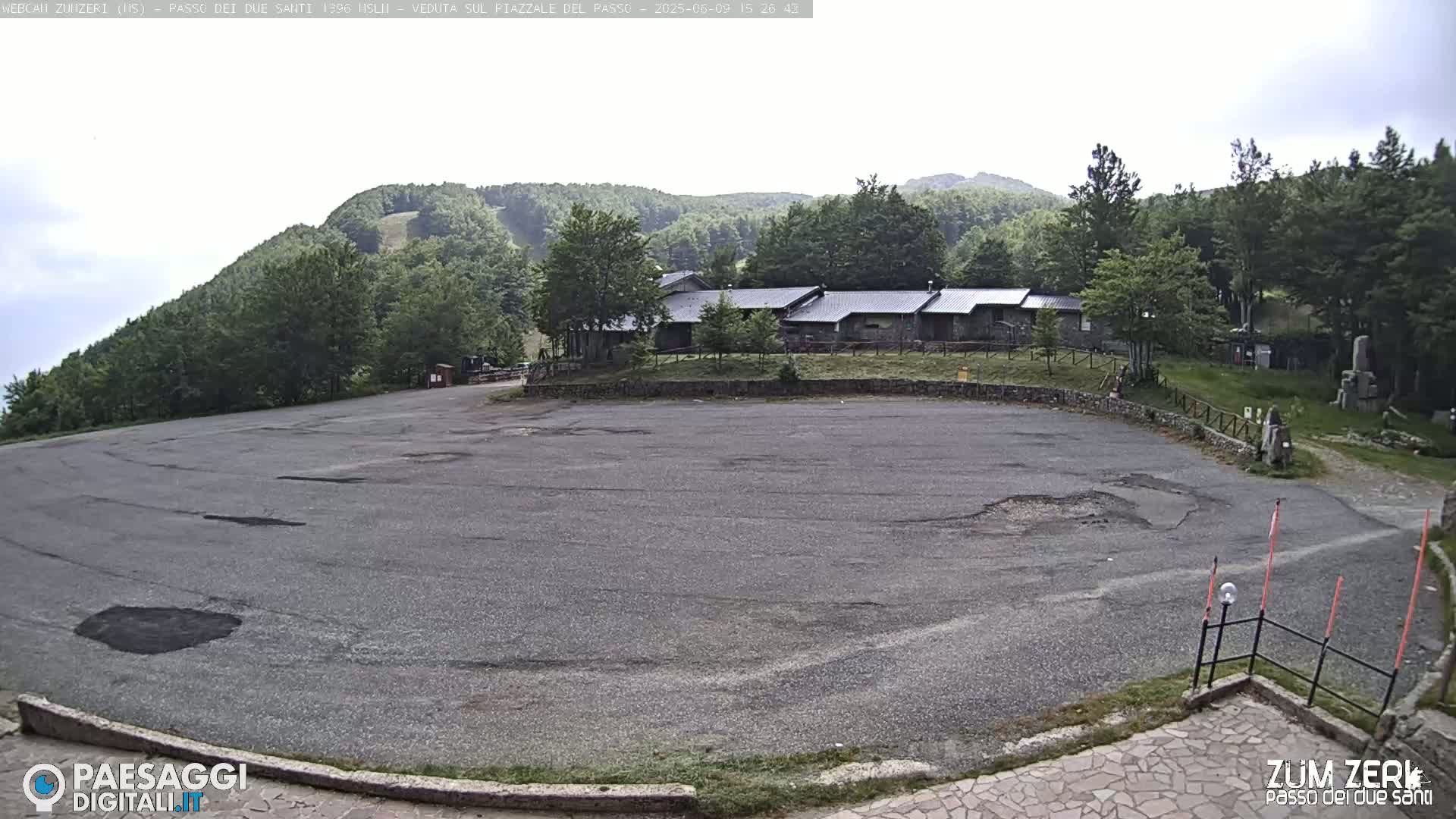 A mostly empty asphalt parking lot sits below a cluster of buildings nestled among green hills under an overcast sky.