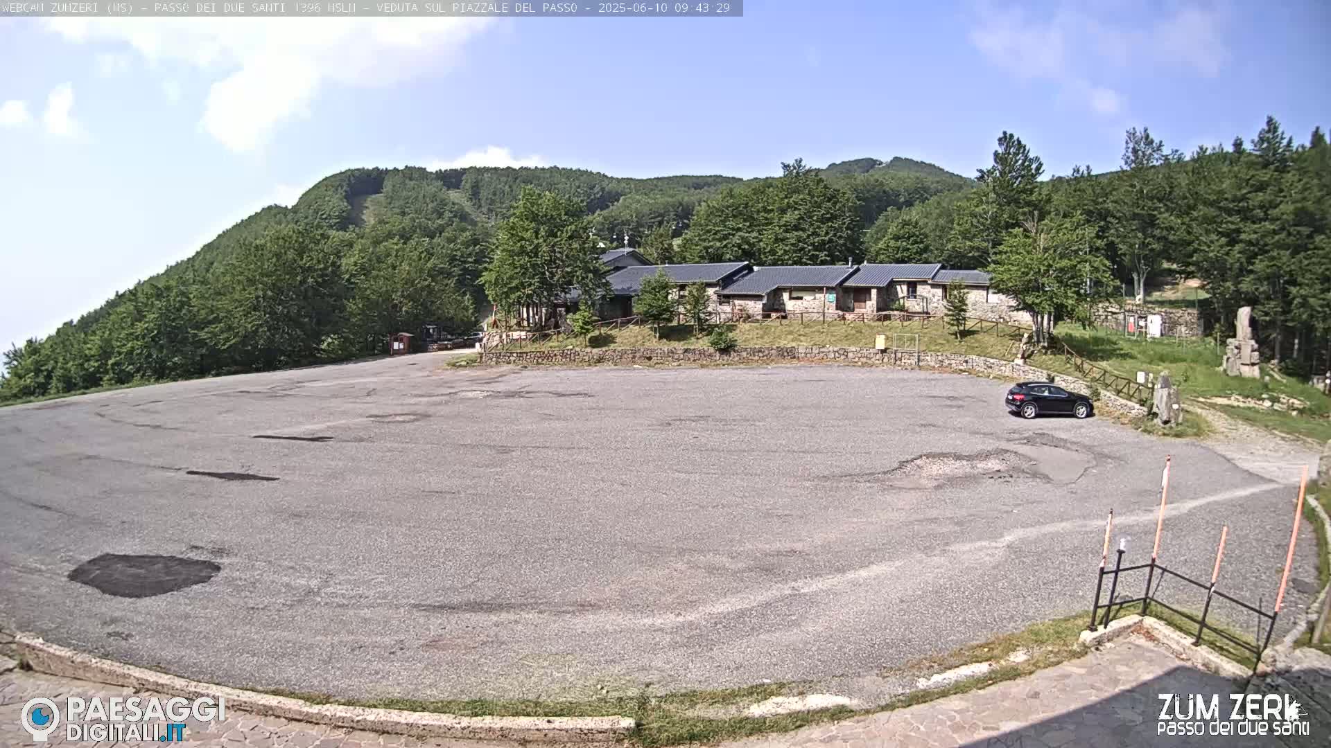 A mostly empty asphalt parking lot sits before a low stone building complex nestled amongst green hills under a clear, sunny sky.