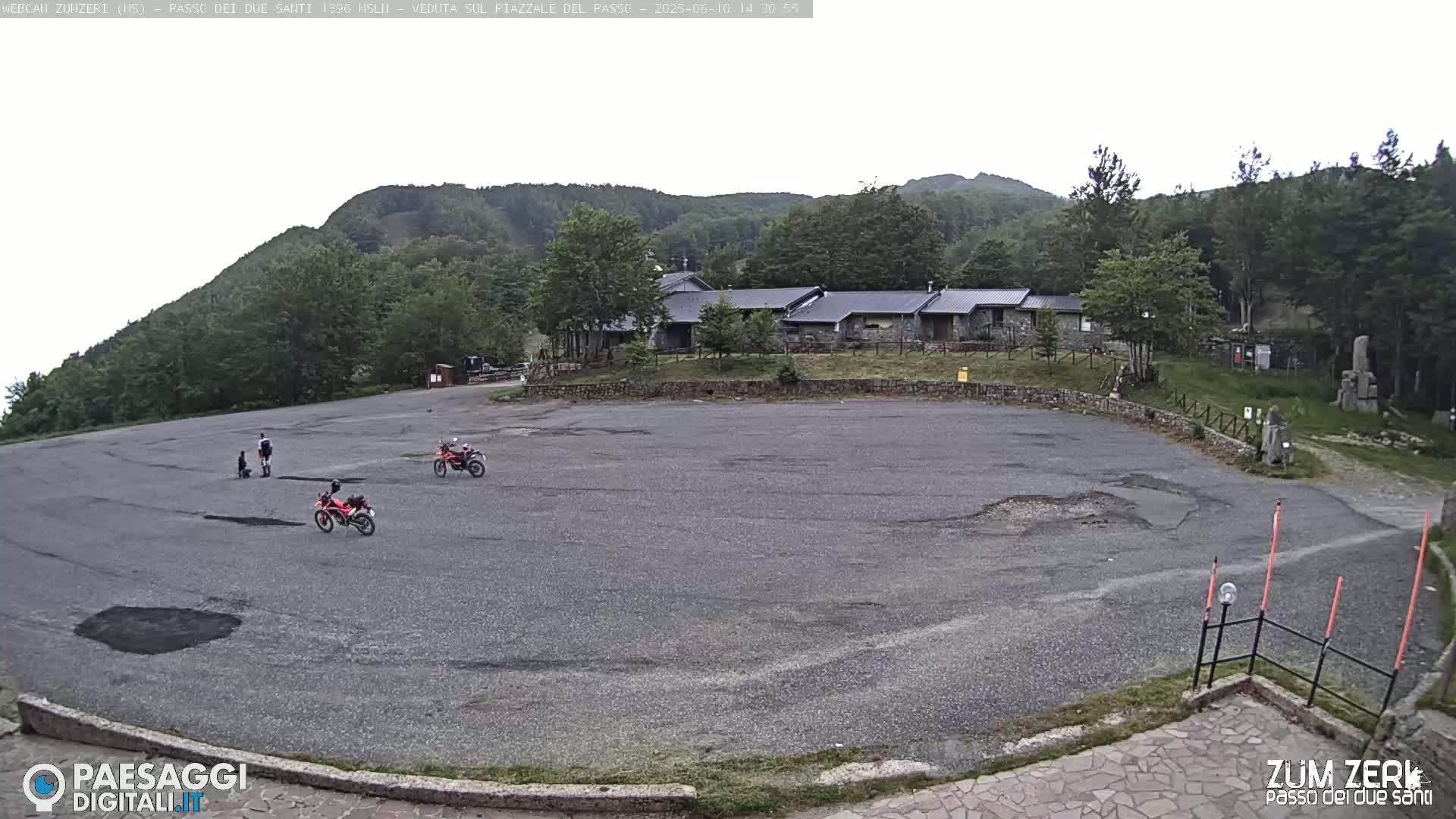 An overcast day reveals a large, empty asphalt parking lot with a few motorcycles and people, situated in front of a stone building and surrounded by green hills and trees.