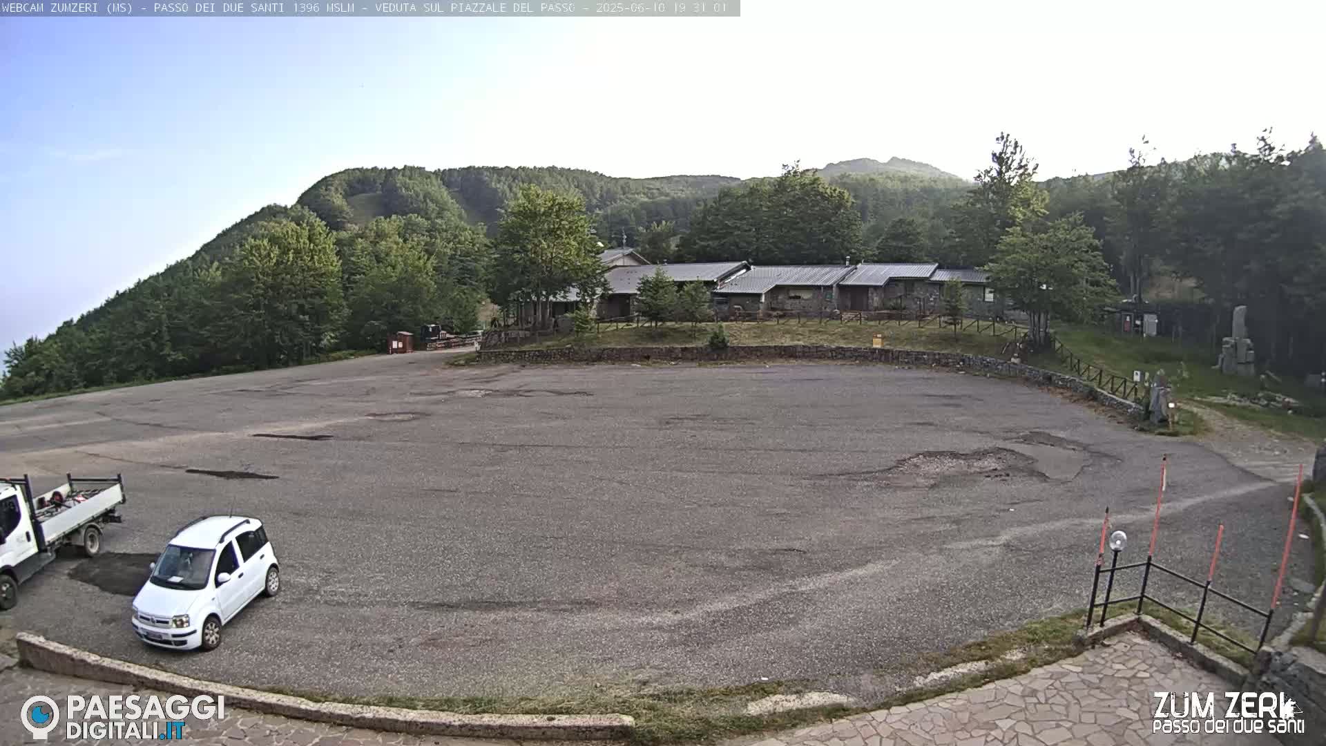 A mostly empty asphalt parking lot sits in front of a cluster of low buildings nestled among forested hills under a mostly clear sky.