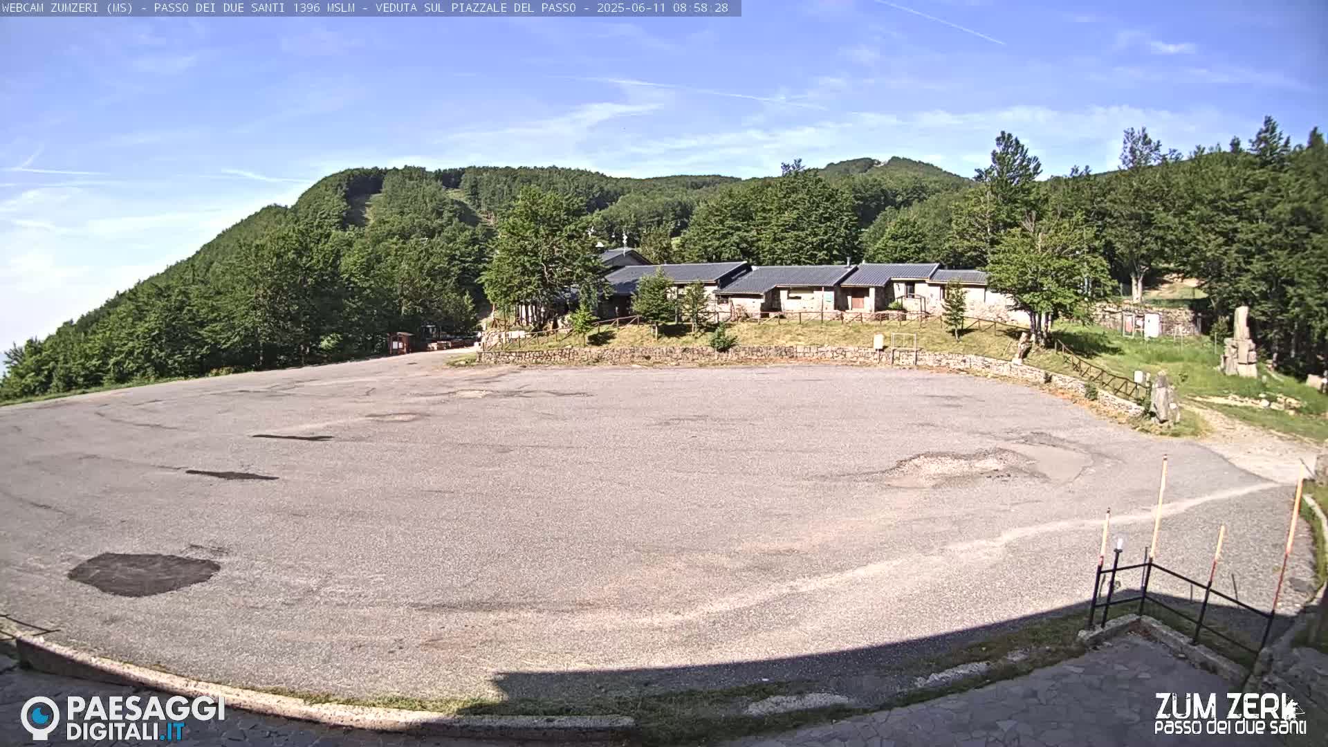 A large, empty asphalt parking lot sits in front of a row of buildings nestled amongst a lush green mountainside under a clear, sunny sky.