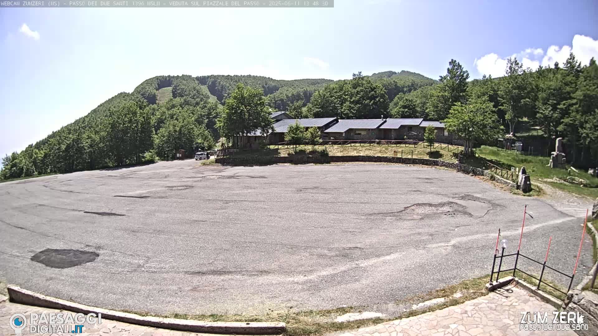 A large, empty asphalt parking lot sits before a cluster of buildings nestled against a verdant hillside under a clear, sunny sky.