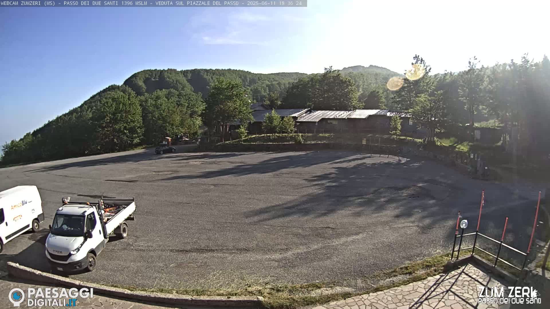 A mostly empty asphalt parking lot sits in front of a cluster of low buildings nestled in a mountainside setting under a clear, sunny sky.