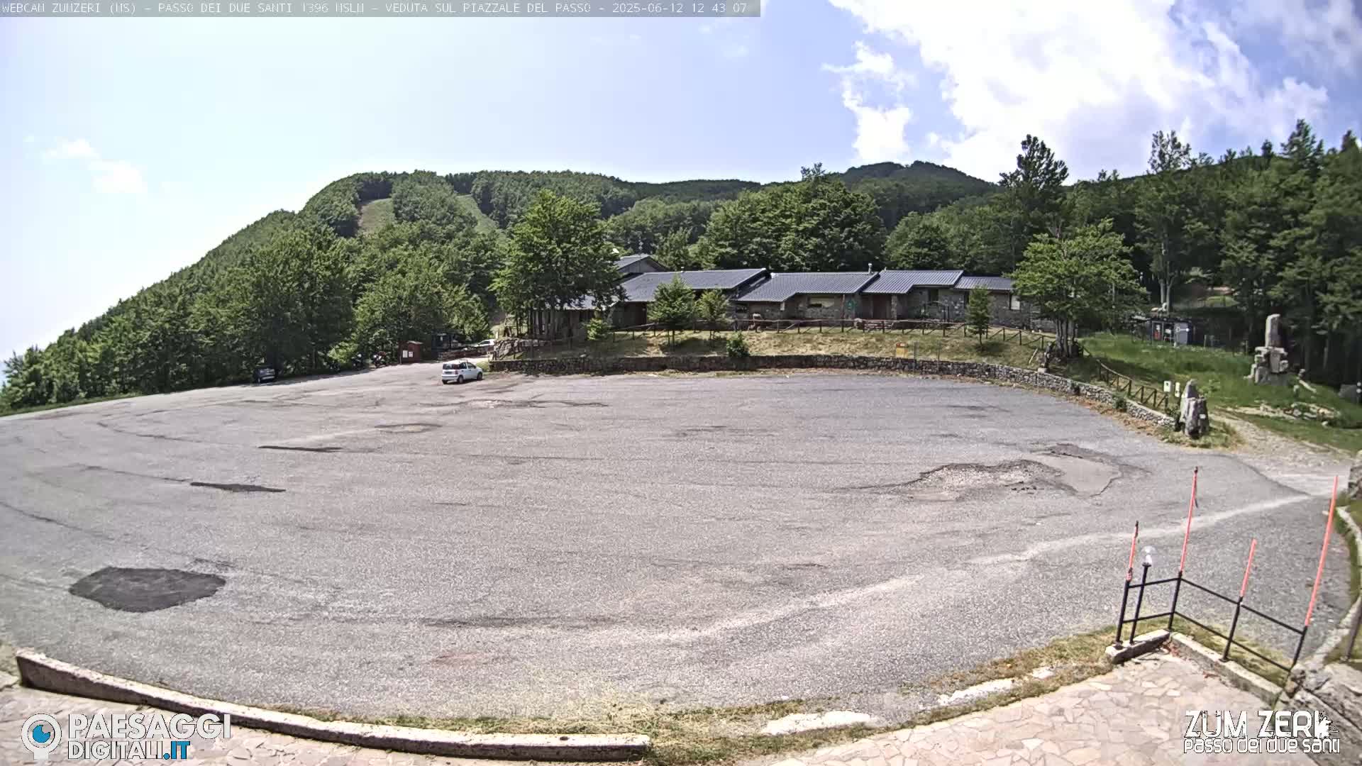 A mostly empty asphalt parking lot sits before a cluster of low buildings, backed by a lush green hillside under a partly sunny sky.