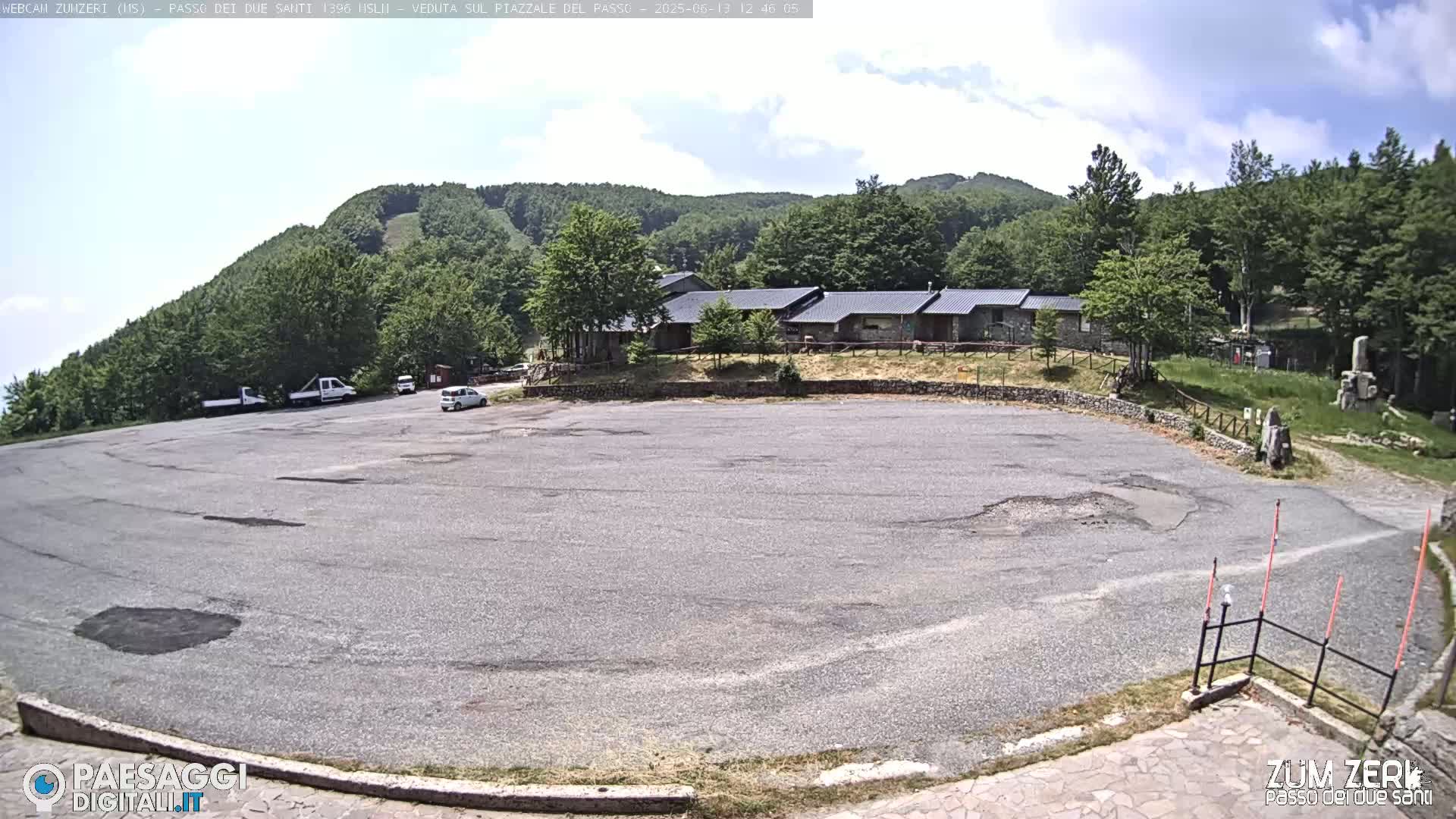 A mostly empty asphalt parking lot sits below a building complex and a forested hillside under a partly cloudy sky.