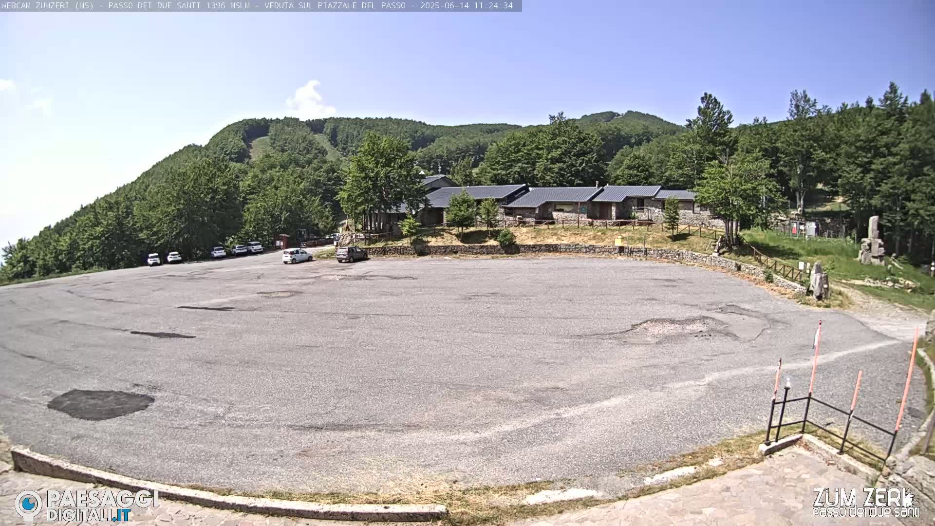 A mostly empty paved parking lot sits below a cluster of buildings nestled amongst a verdant hillside under a clear blue sky.