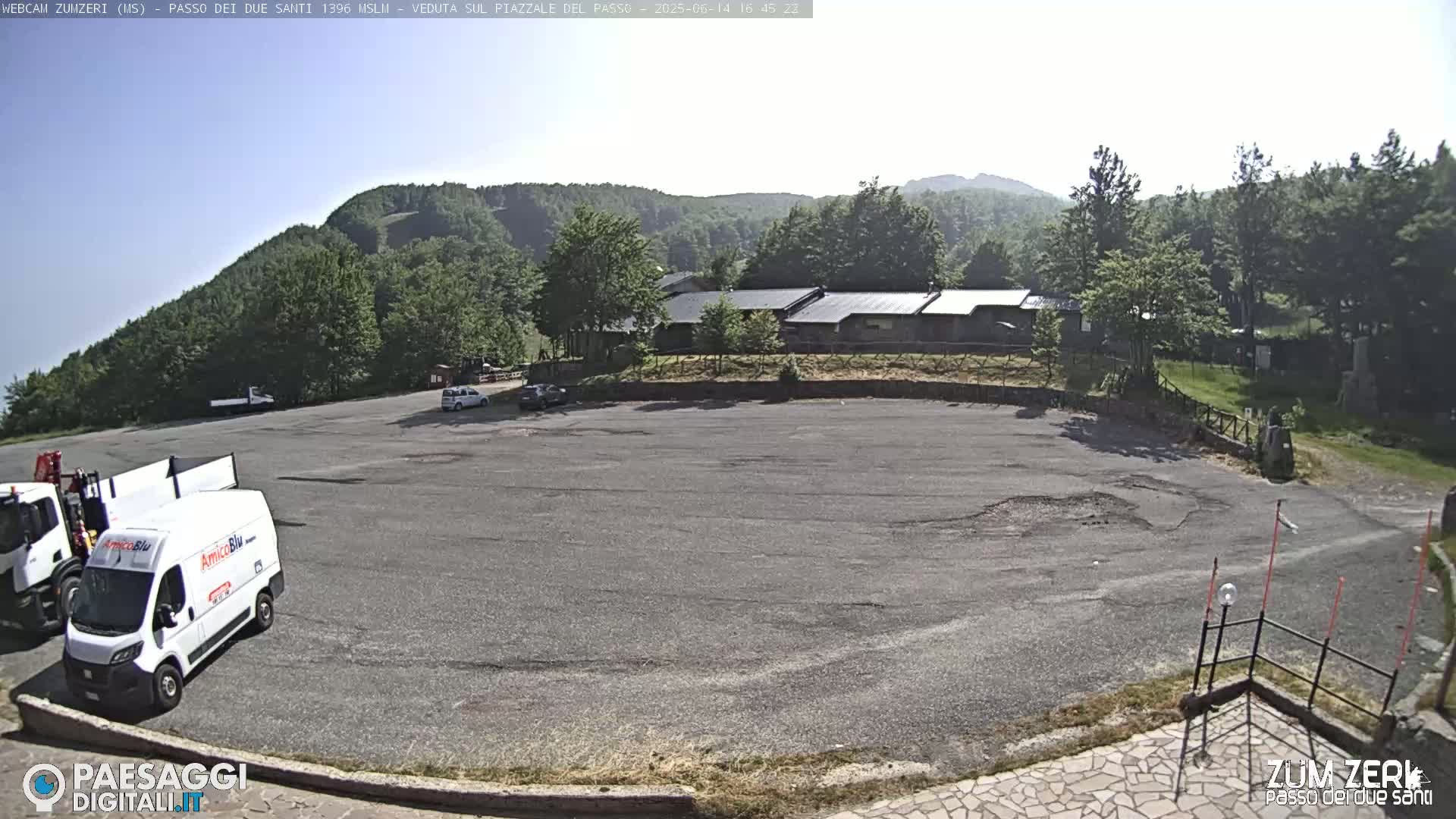 A large, mostly empty asphalt parking lot is situated in front of a building complex nestled within a mountainous, wooded area under a mostly clear sky.