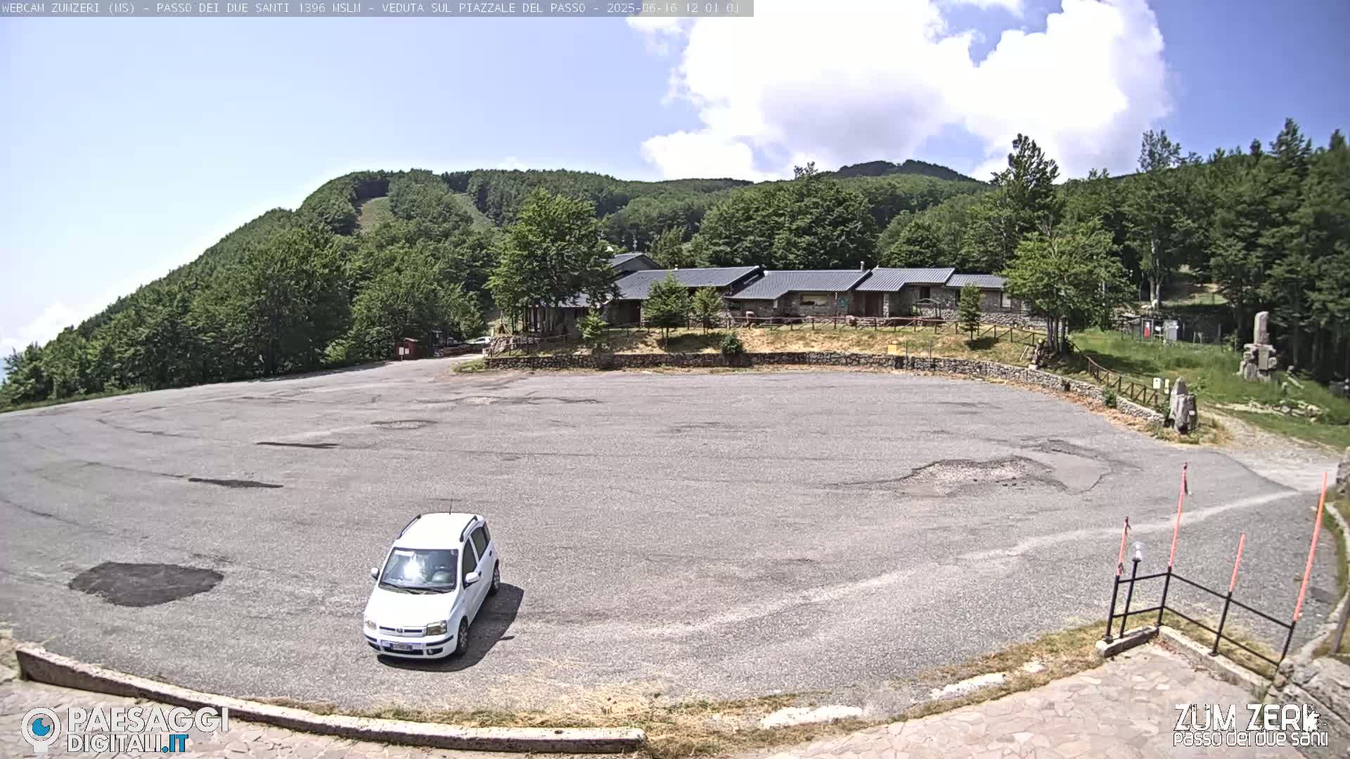 A mostly empty asphalt parking lot sits before a complex of buildings nestled amongst a lush green hillside under a partly cloudy sky.