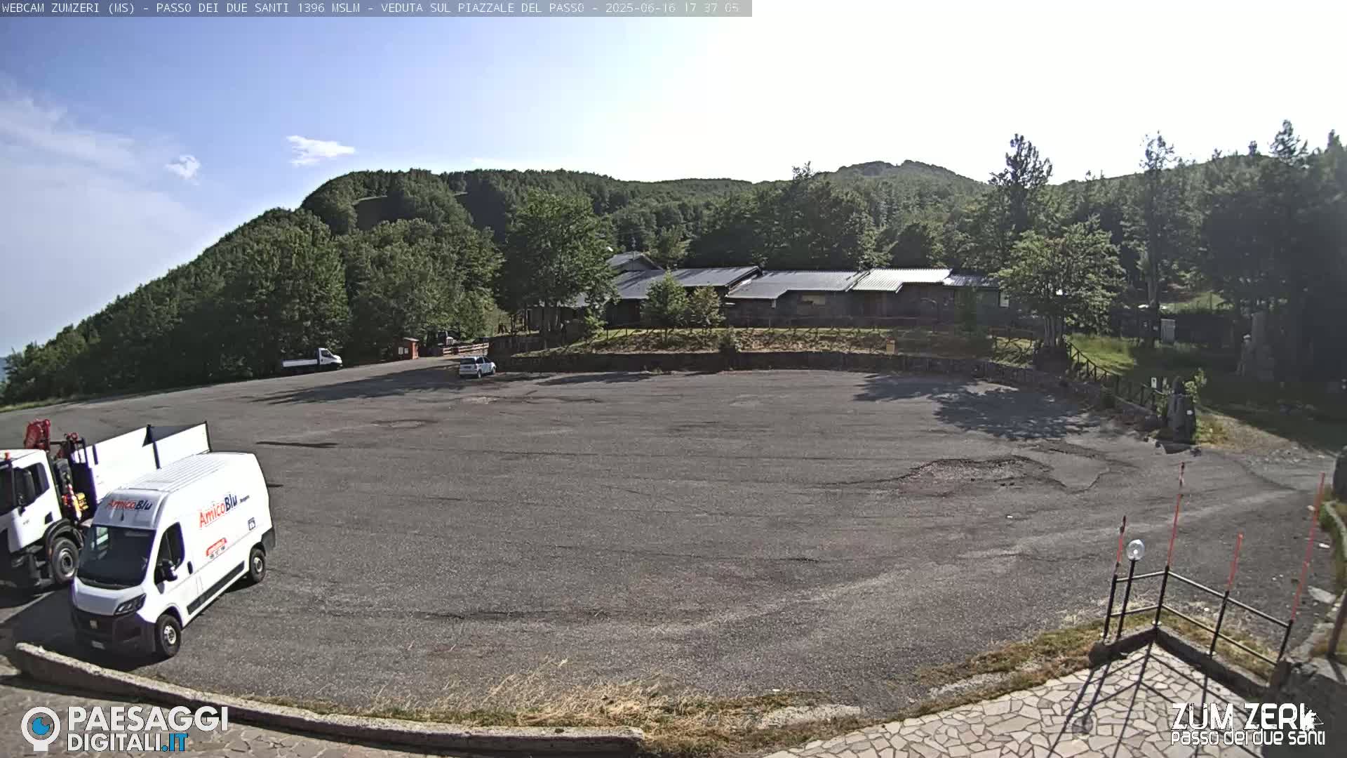 A mostly empty asphalt parking lot sits before a complex of low buildings nestled against a tree-covered hillside under a mostly clear sky.