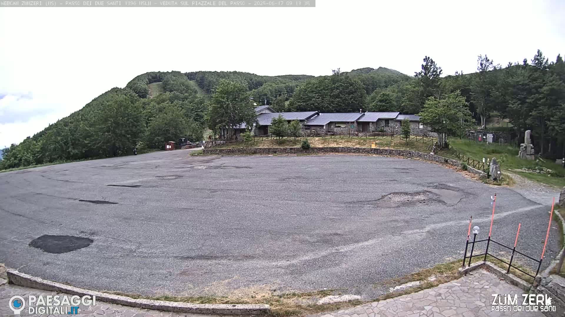 A large, empty asphalt parking lot with potholes is situated in front of a stone building complex nestled among green hills under an overcast sky.