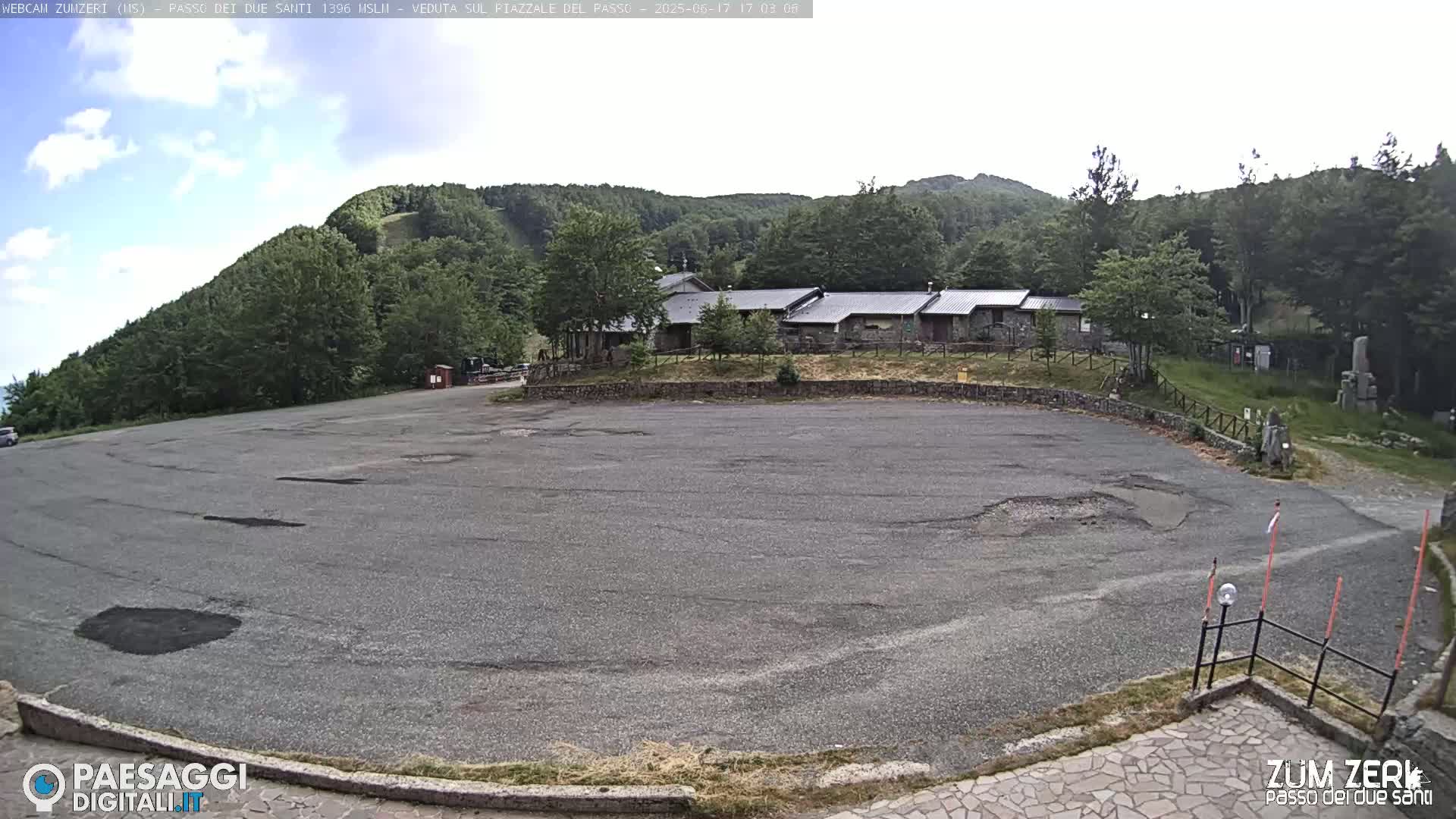 A mostly empty asphalt parking lot sits before a low stone building nestled amongst a lush green, hilly landscape under a partly cloudy sky.