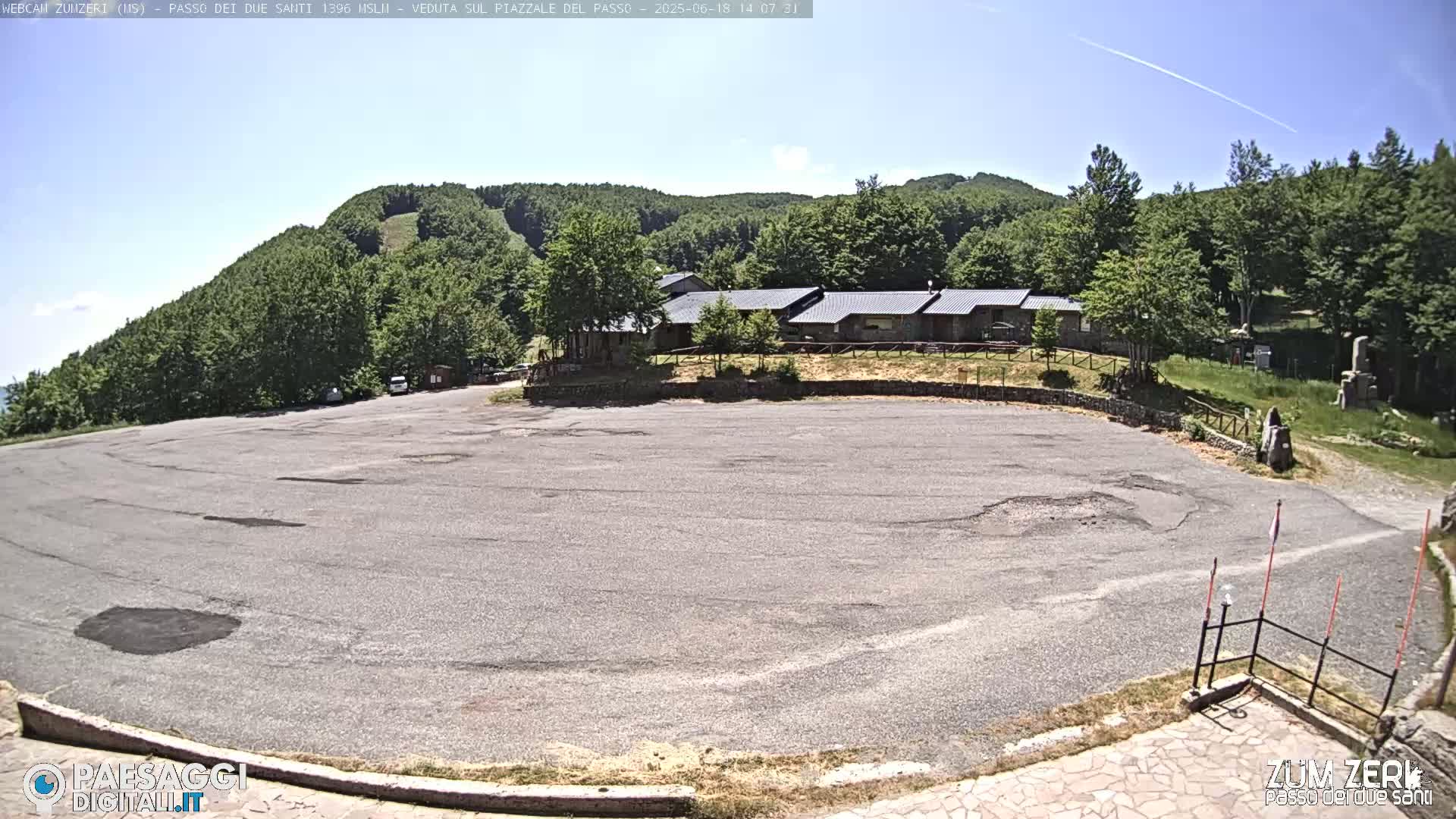 An empty asphalt parking lot is situated in front of a low building complex, nestled amongst a lush green, hilly landscape under a clear, sunny sky.