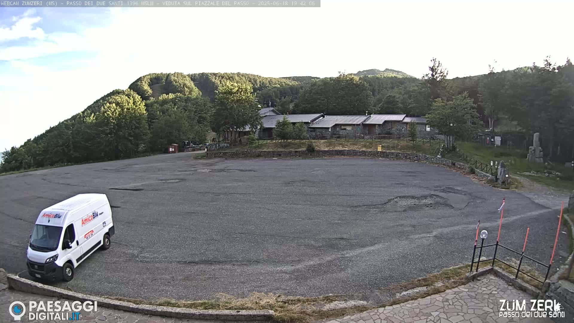 A mostly empty asphalt parking lot sits in front of a cluster of low buildings, nestled amongst a dark green, forested hillside under a mostly clear sky.