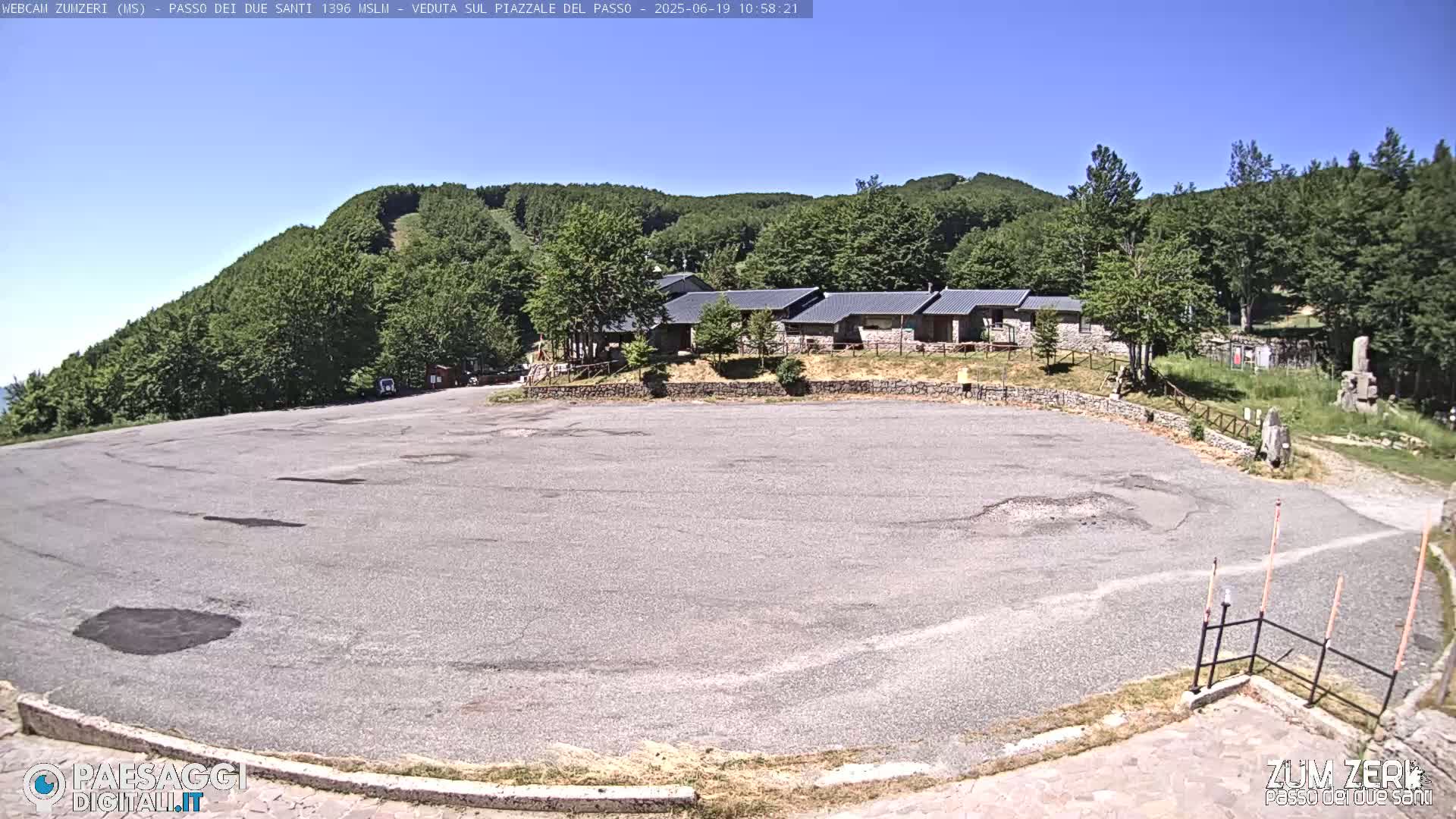 A large, empty asphalt parking lot sits in front of a cluster of low buildings, all nestled amongst a lush green hillside under a clear blue sky.