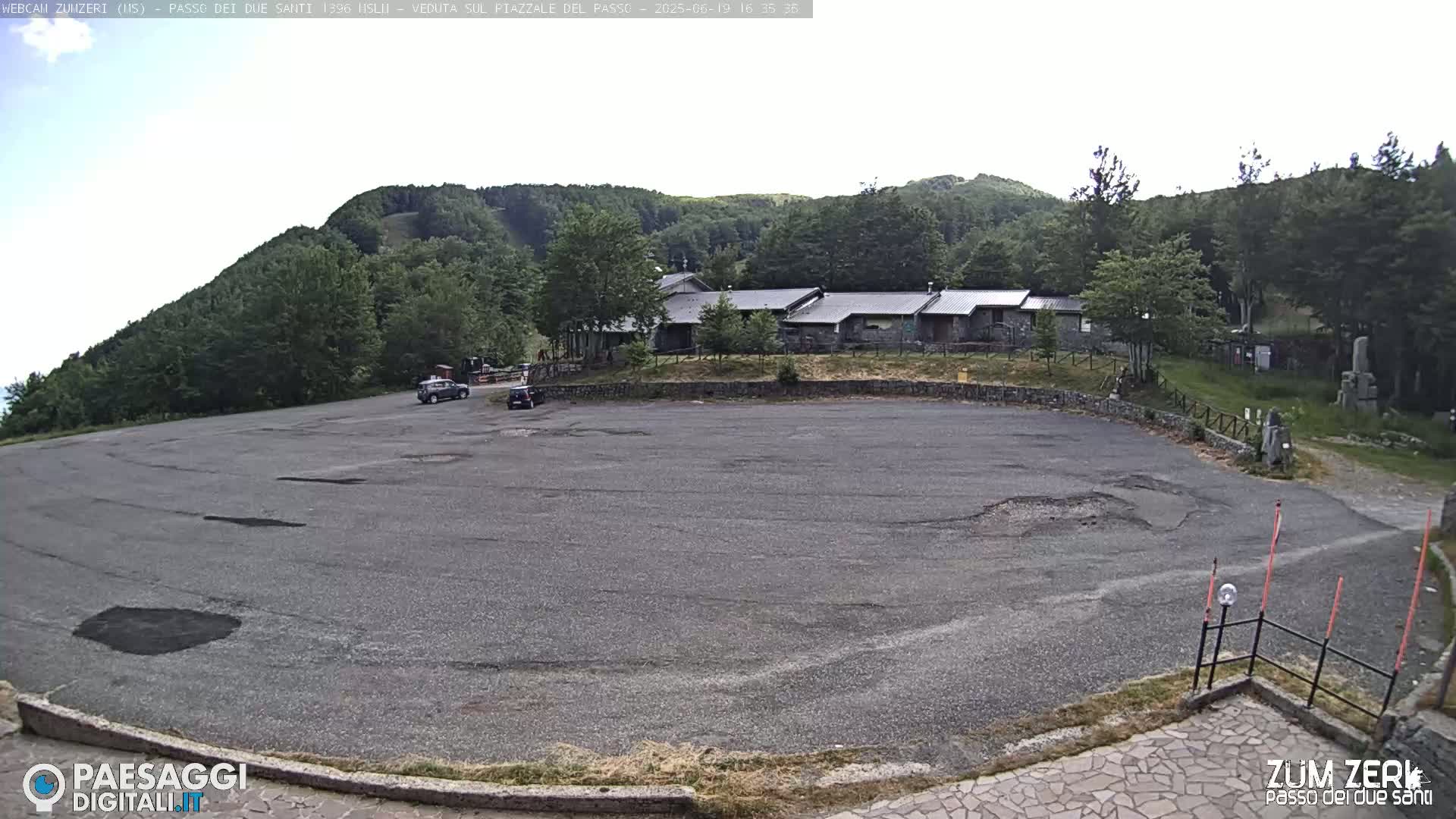 A mostly empty asphalt parking lot is situated in front of a cluster of buildings nestled among a lush green hillside under a bright, overcast sky.