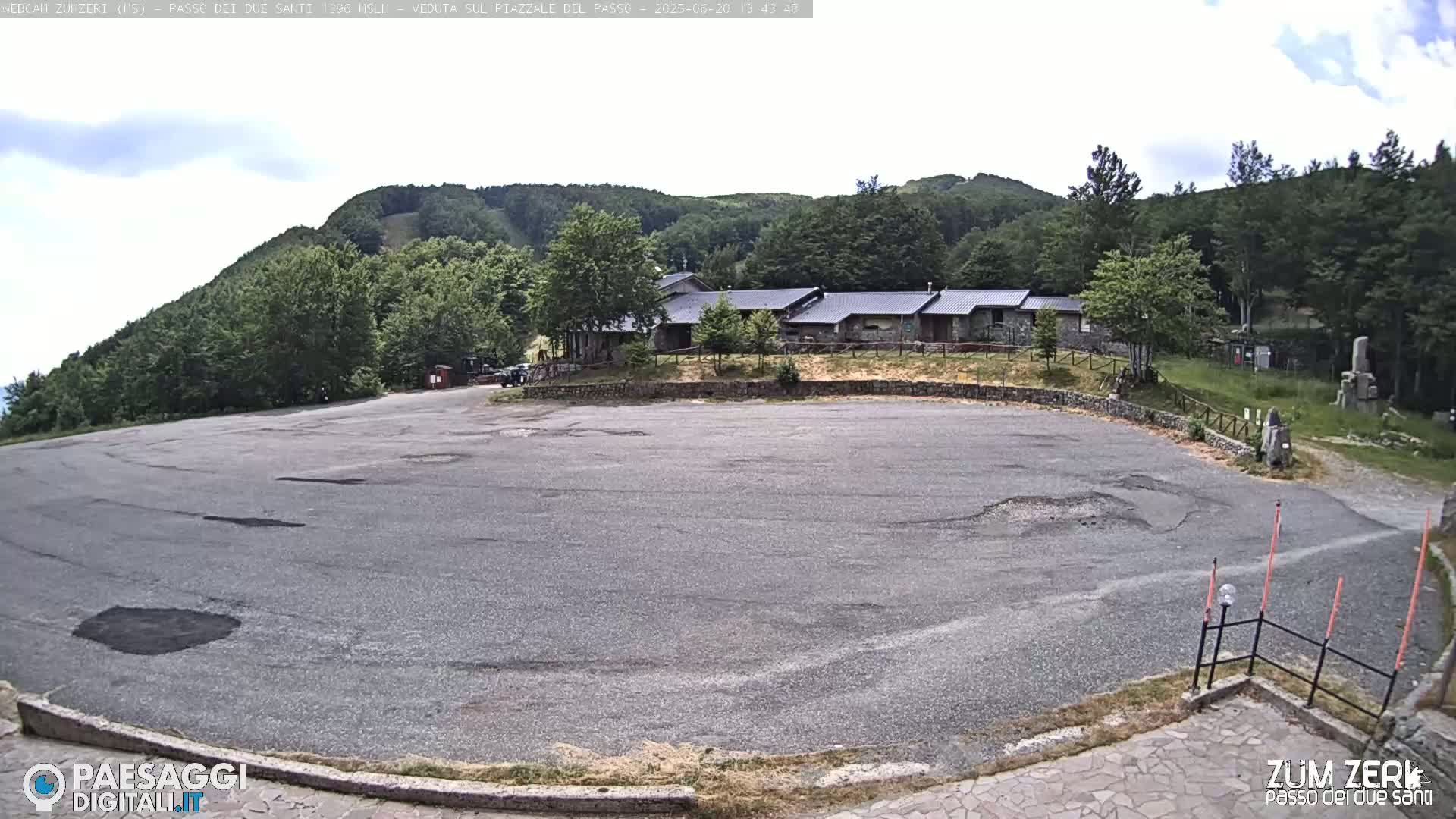 An empty asphalt parking lot sits before a cluster of buildings nestled among a verdant, hilly landscape under a partly cloudy sky.