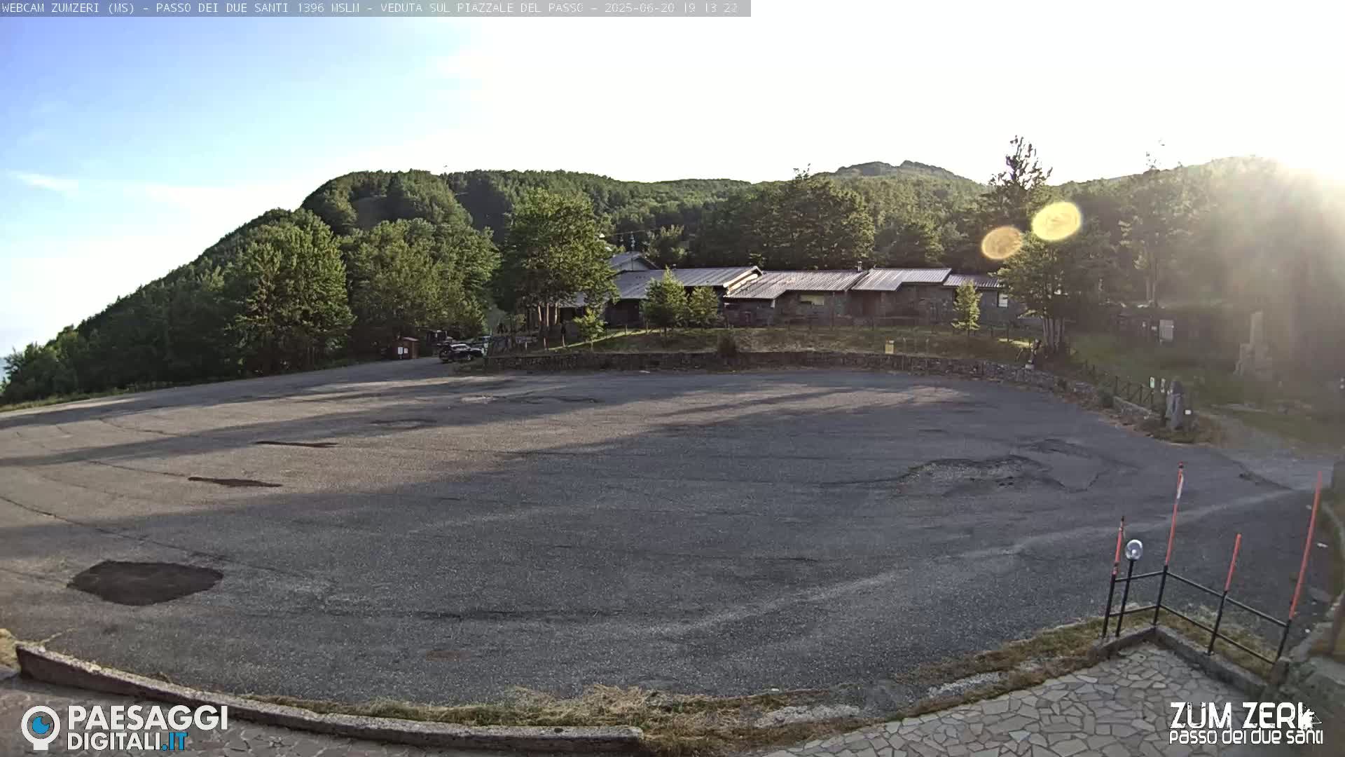 A mostly empty asphalt parking lot sits below a cluster of buildings nestled in a forest on a sunny day.