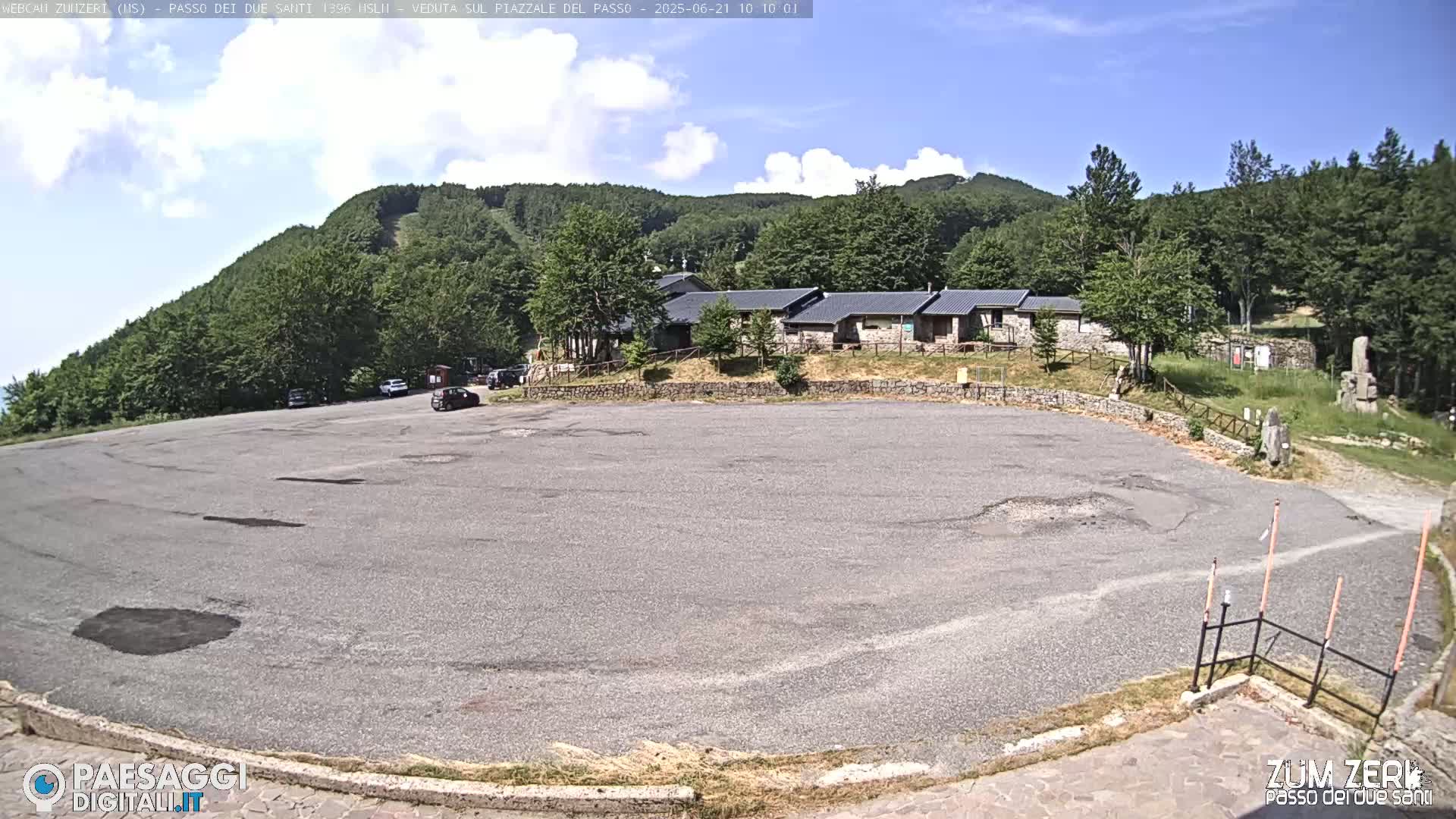 A mostly empty asphalt parking lot sits before a low stone building complex nestled amongst a lush green hillside under a partly cloudy sky.