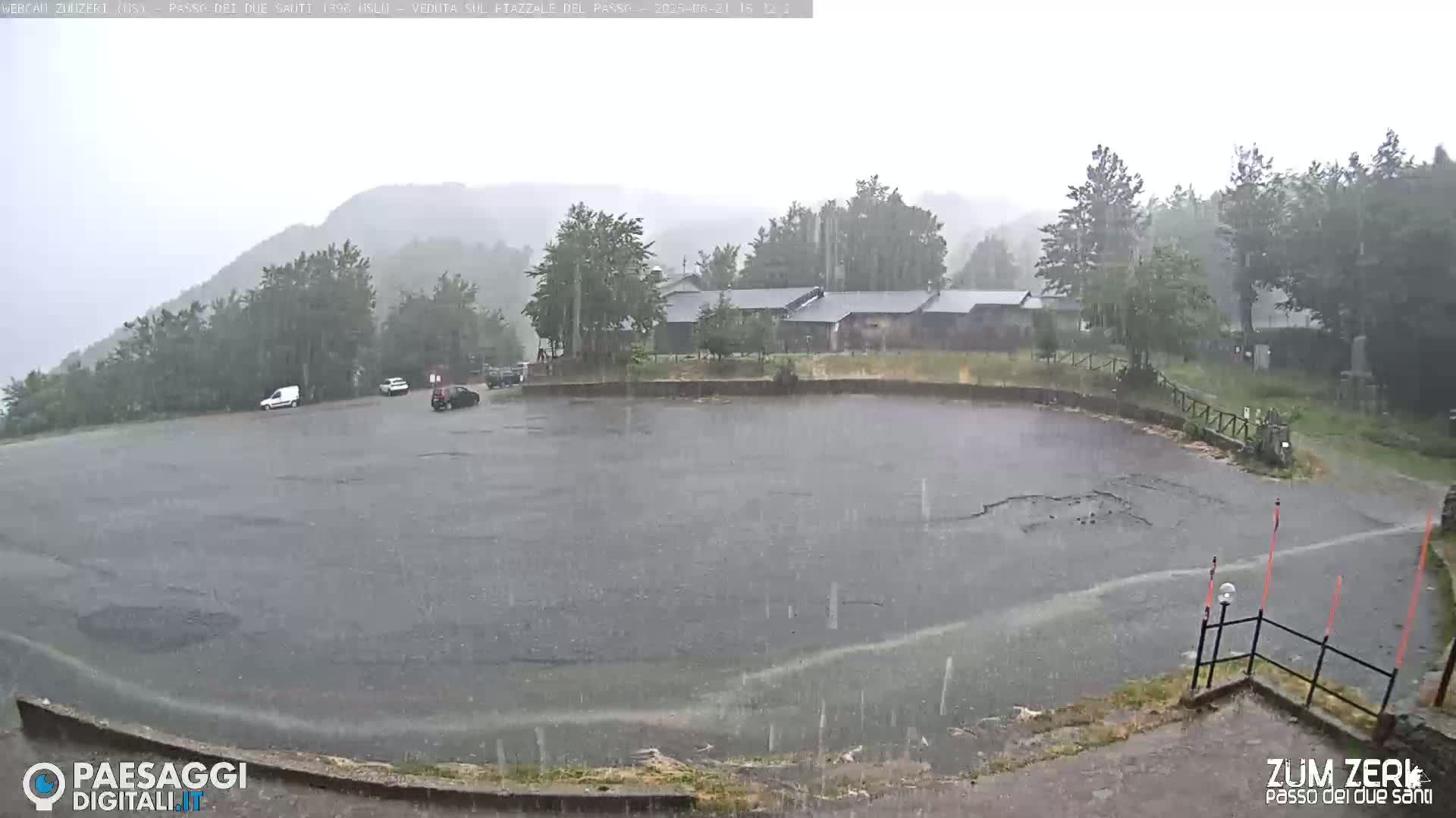 A rain-soaked empty parking lot is seen, with a few vehicles parked and buildings in the background under heavy rainfall.