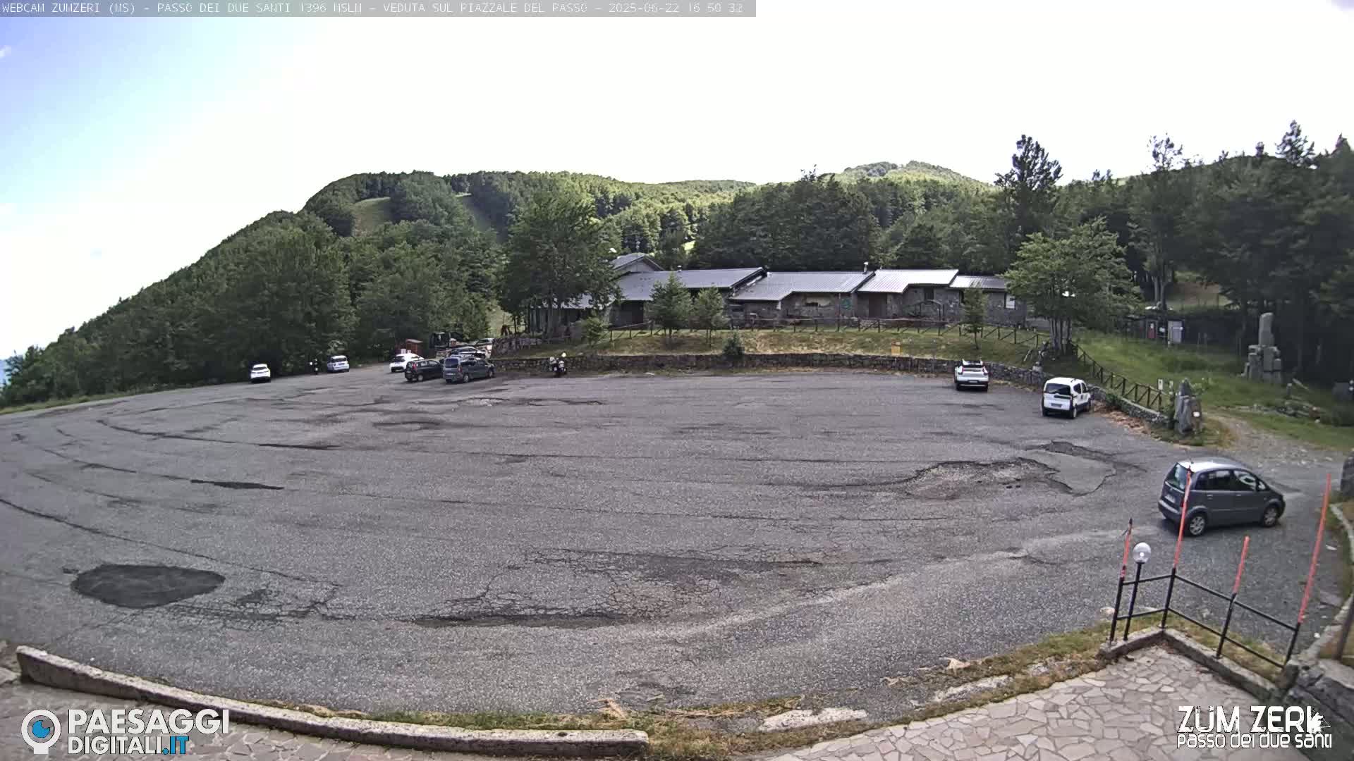 A mostly empty asphalt parking lot with a few cars sits in front of a stone building nestled amongst a green, forested hillside under a partly cloudy sky.
